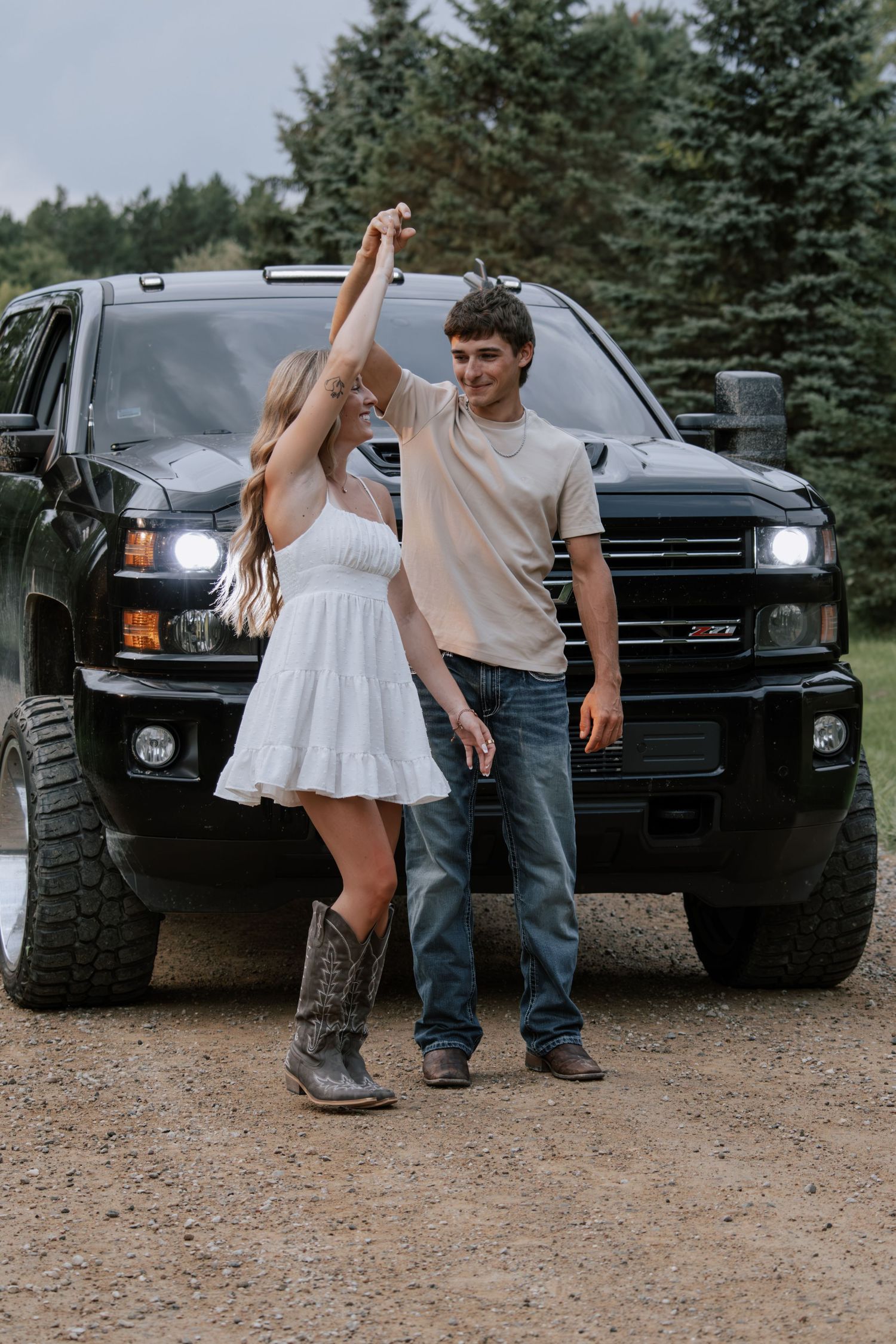 A young couple dances together in front of a black lifted truck on a gravel path surrounded by evergreen trees.
