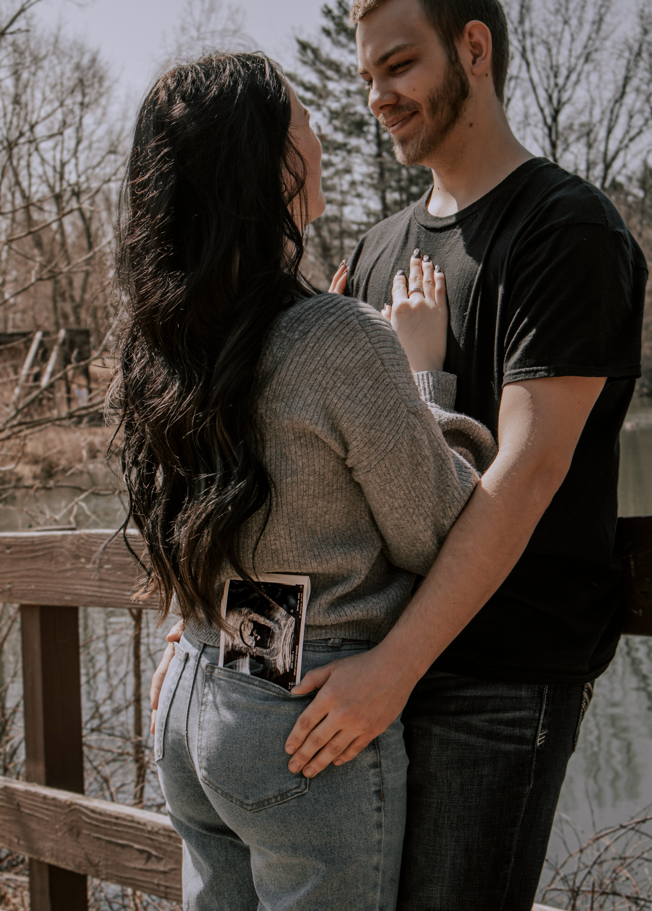 Couple embracing on a bridge holding an ultrasound photo, announcing pregnancy outdoors near a serene waterway.