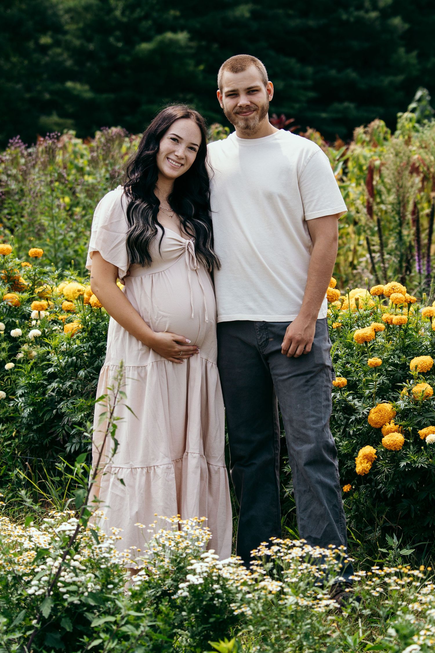 Smiling pregnant woman in blush maxi dress poses with partner in white tee among yellow marigolds in lush garden.