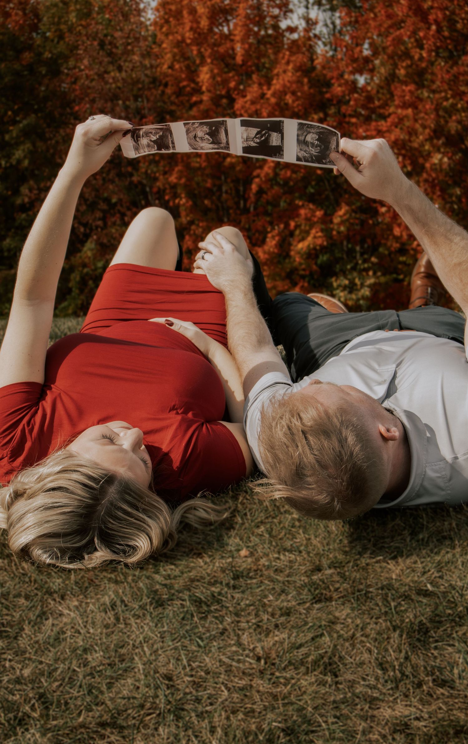 Couple lying on autumn grass with vibrant orange foliage in the background during a fall photoshoot.