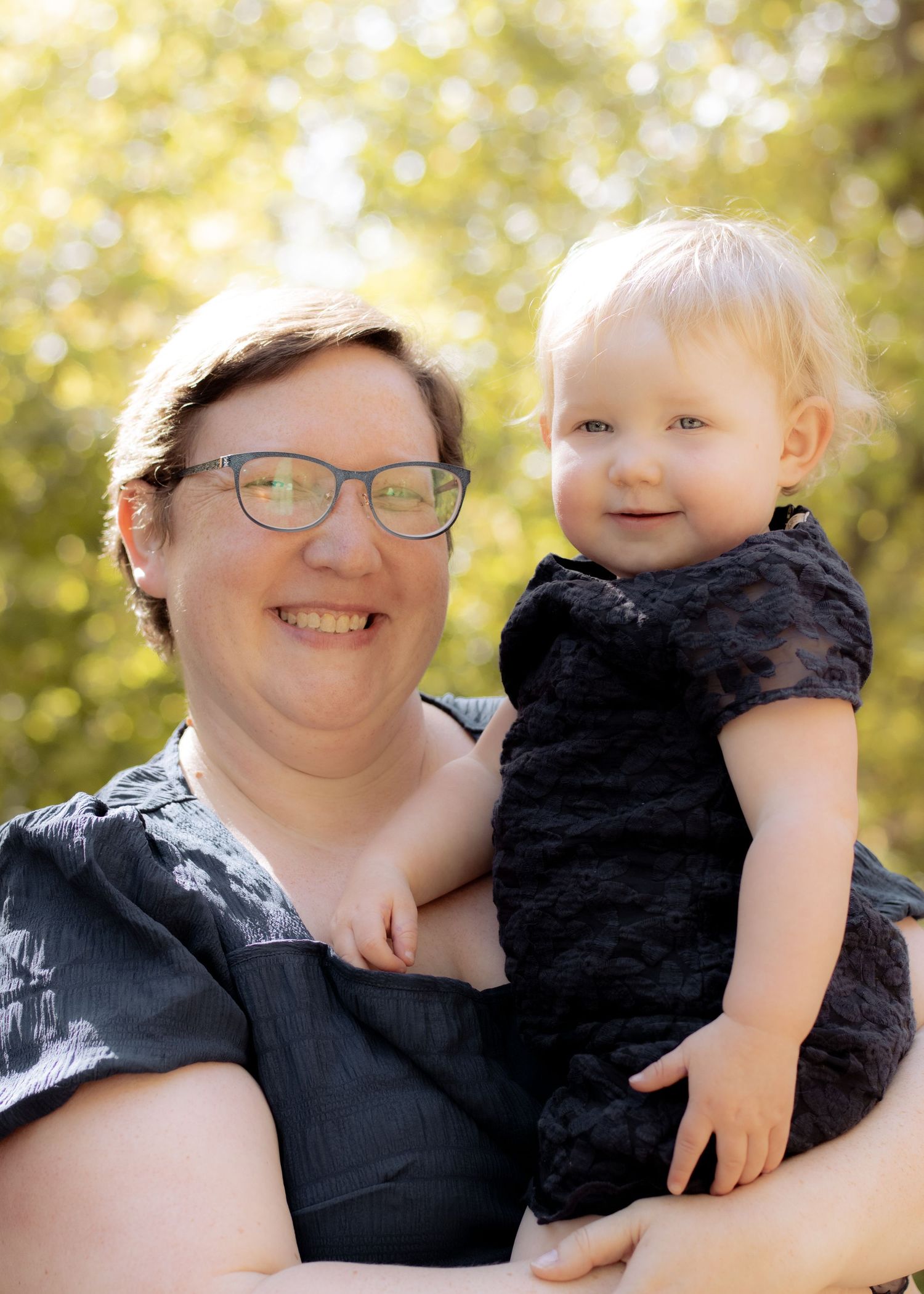 Smiling woman with glasses holds a happy blonde toddler outdoors in soft natural light.