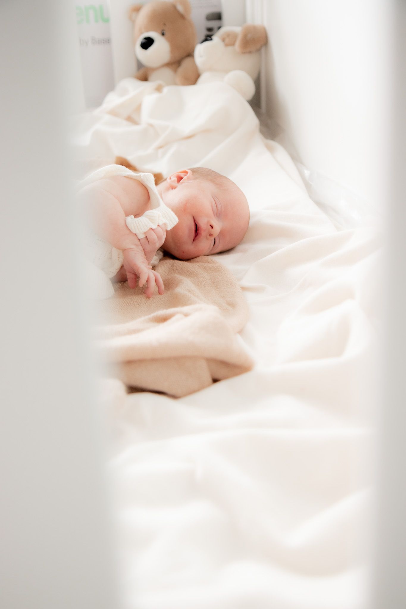 Peaceful newborn baby sleeping in white crib with teddy bear and soft white blanket.