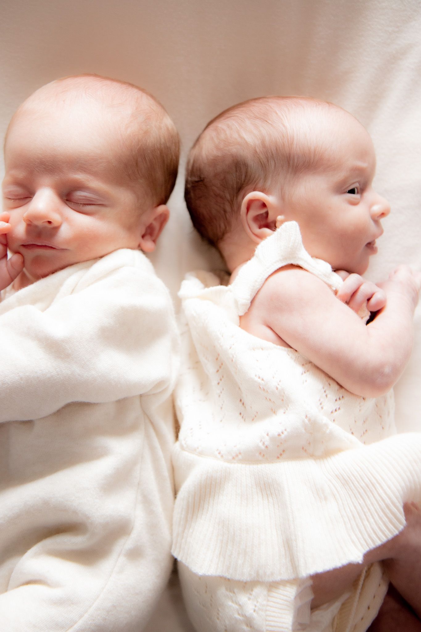 Two newborn twins lying back to back in white knit outfits on a soft cream blanket.