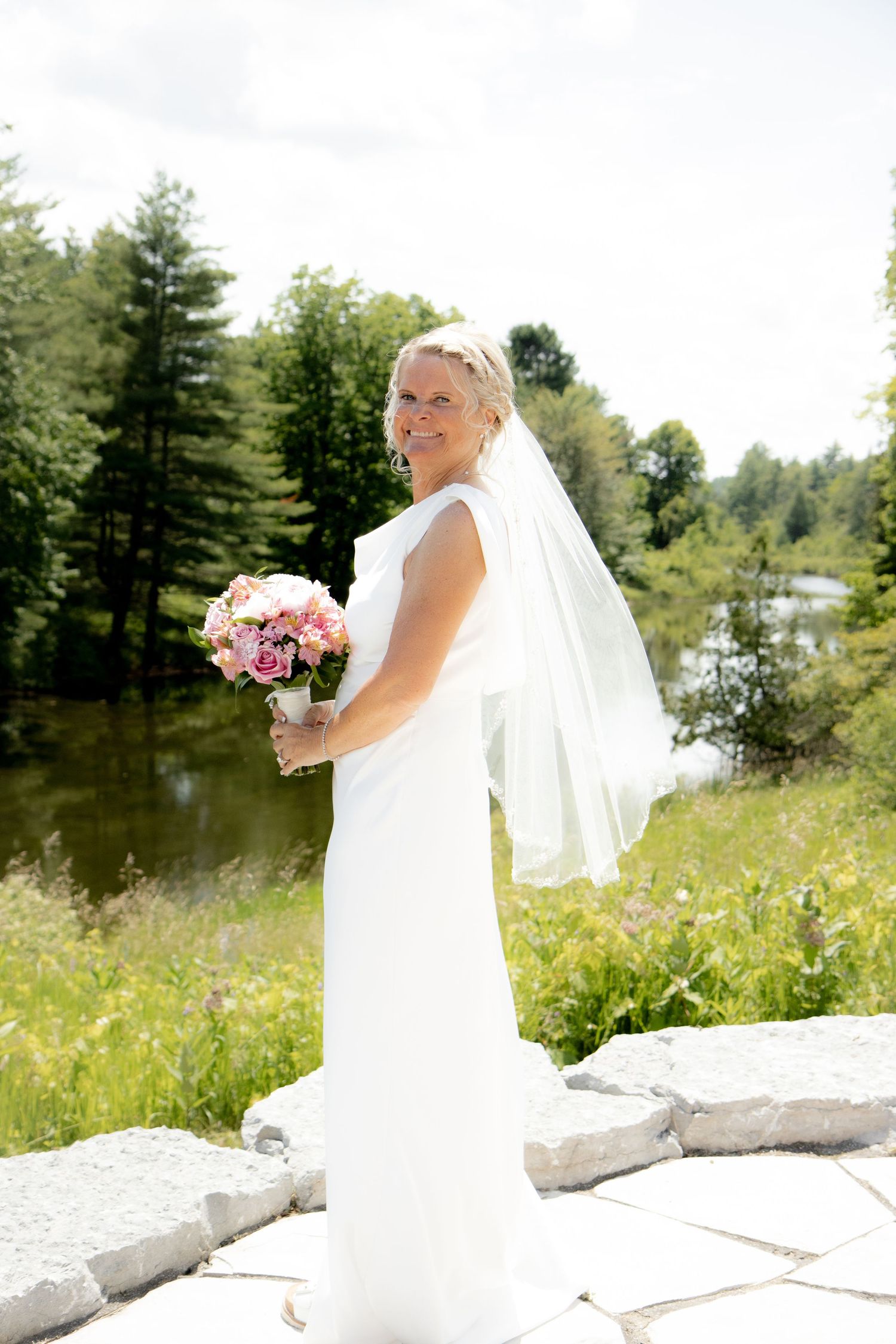 Smiling bride in white gown and veil holds pink bouquet outdoors near a serene lake surrounded by lush green trees.