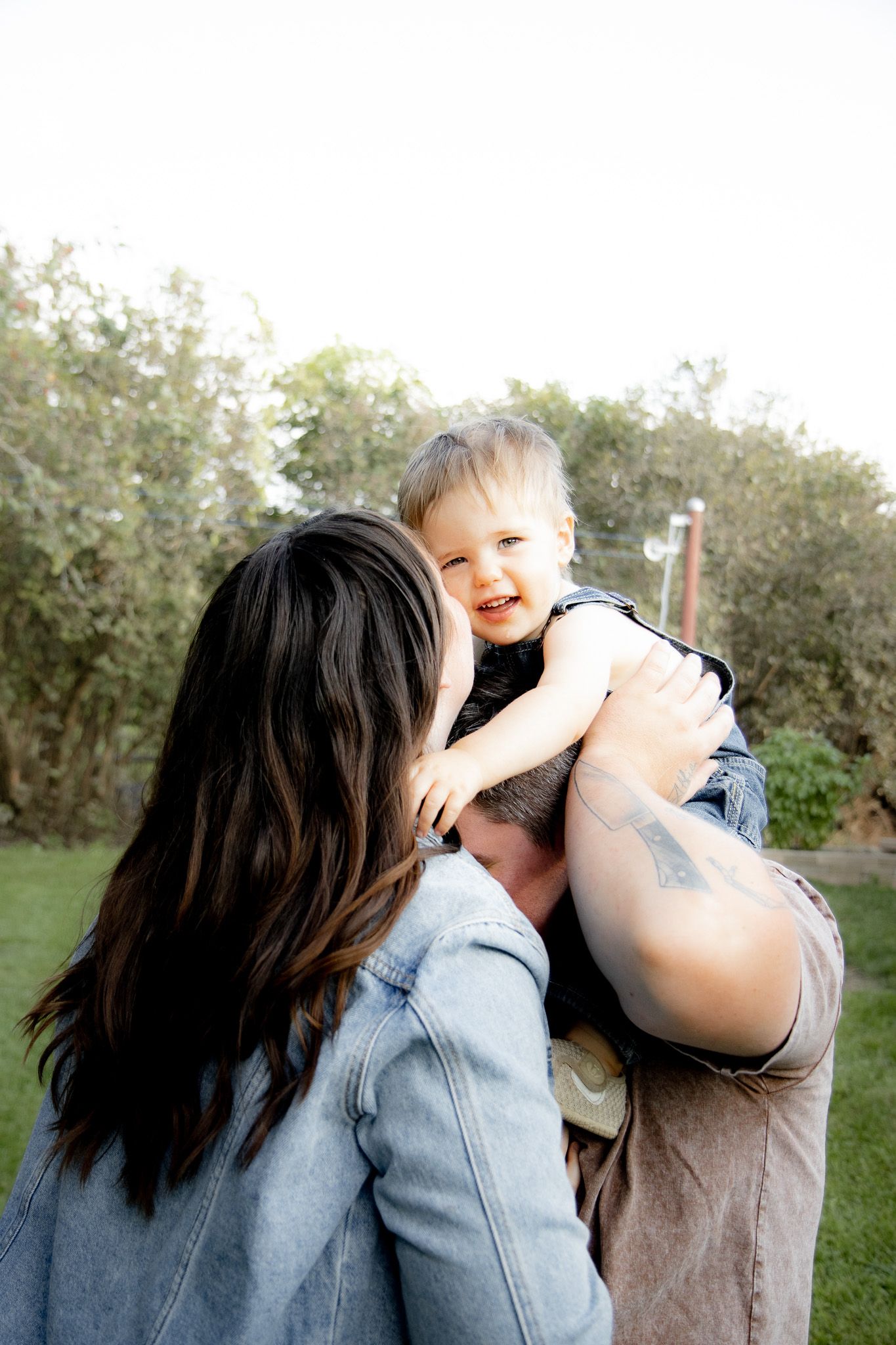 A woman with dark hair holds a laughing baby outdoors, both enjoying a joyful moment in a green garden.
