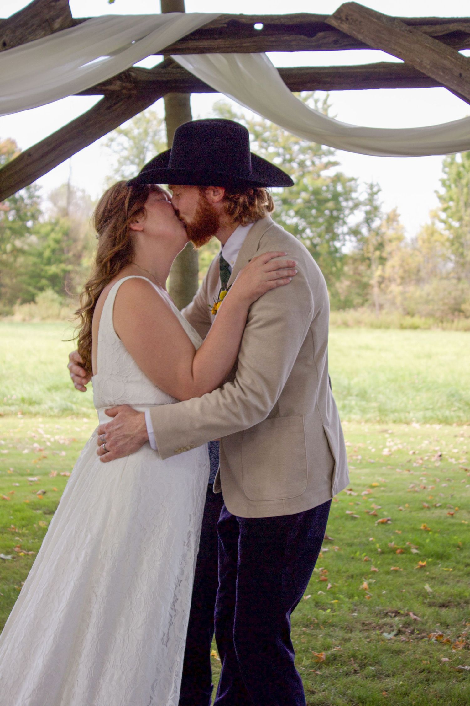 Bride and groom kiss under rustic wooden arch during outdoor fall wedding ceremony.