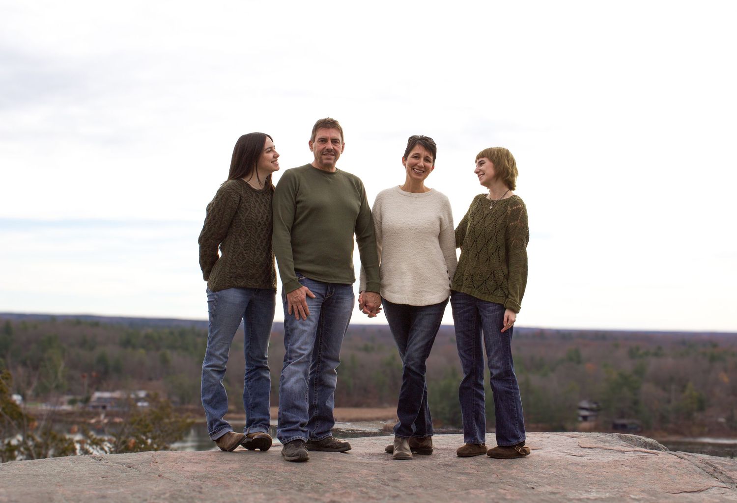 Family of four stands holding hands on rocky hilltop with vast autumn landscape stretching behind them.
