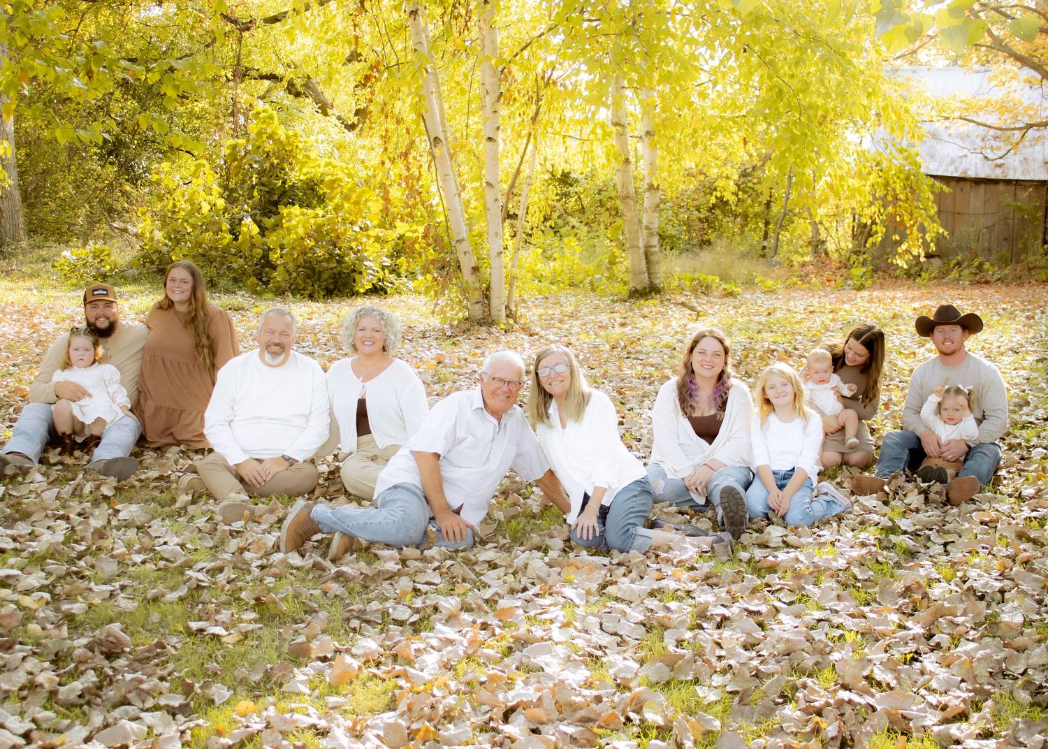 Large multigenerational family sits together on fallen leaves beneath golden autumn trees during outdoor photo session.