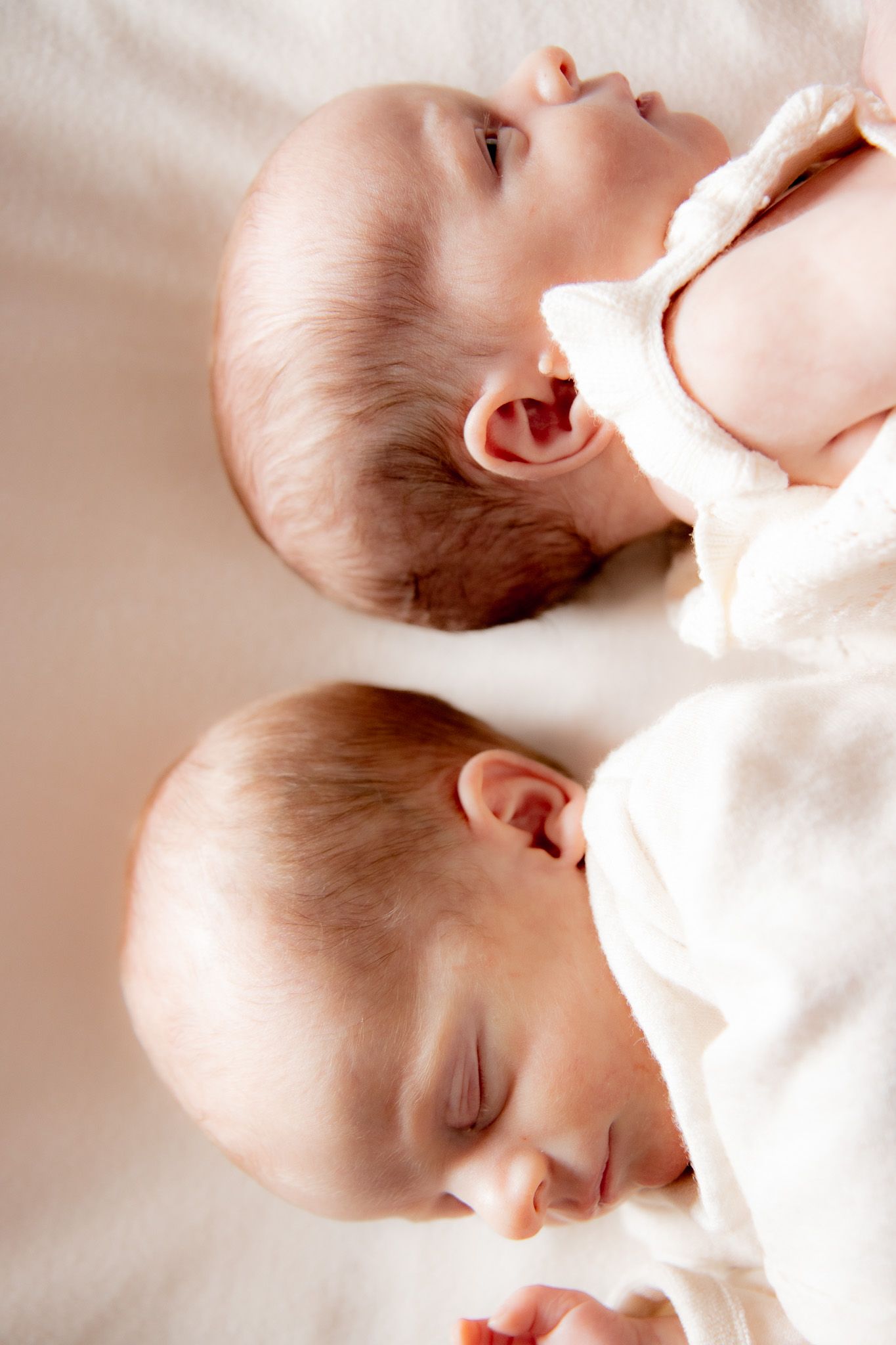 Two newborn twins lying side by side on a white blanket, one sleeping and one awake.