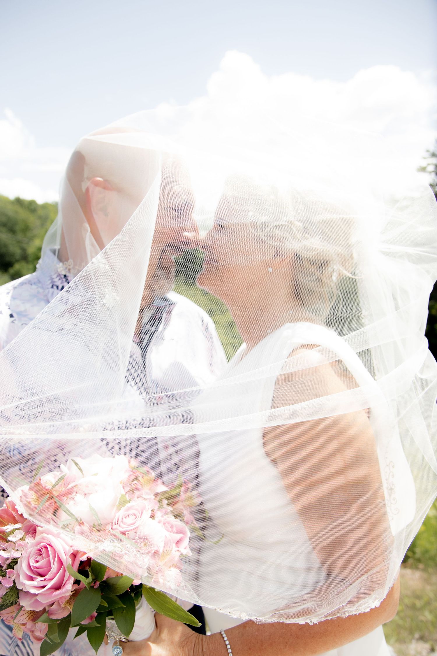 Two brides sharing a kiss under a flowing white veil, holding pink rose bouquet on their wedding day.