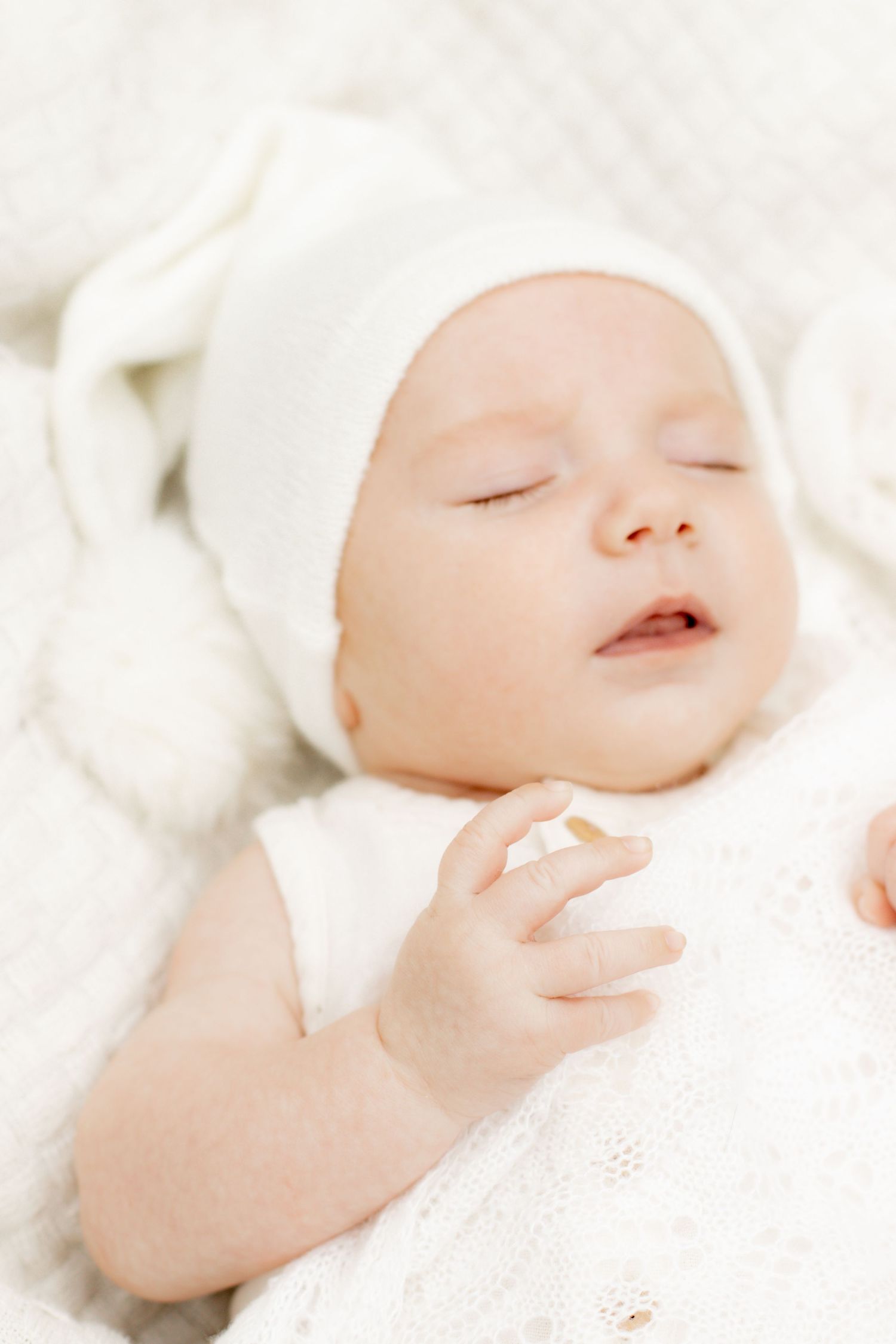 Adorable sleeping baby in a white knit hat lying on a soft white lace blanket.