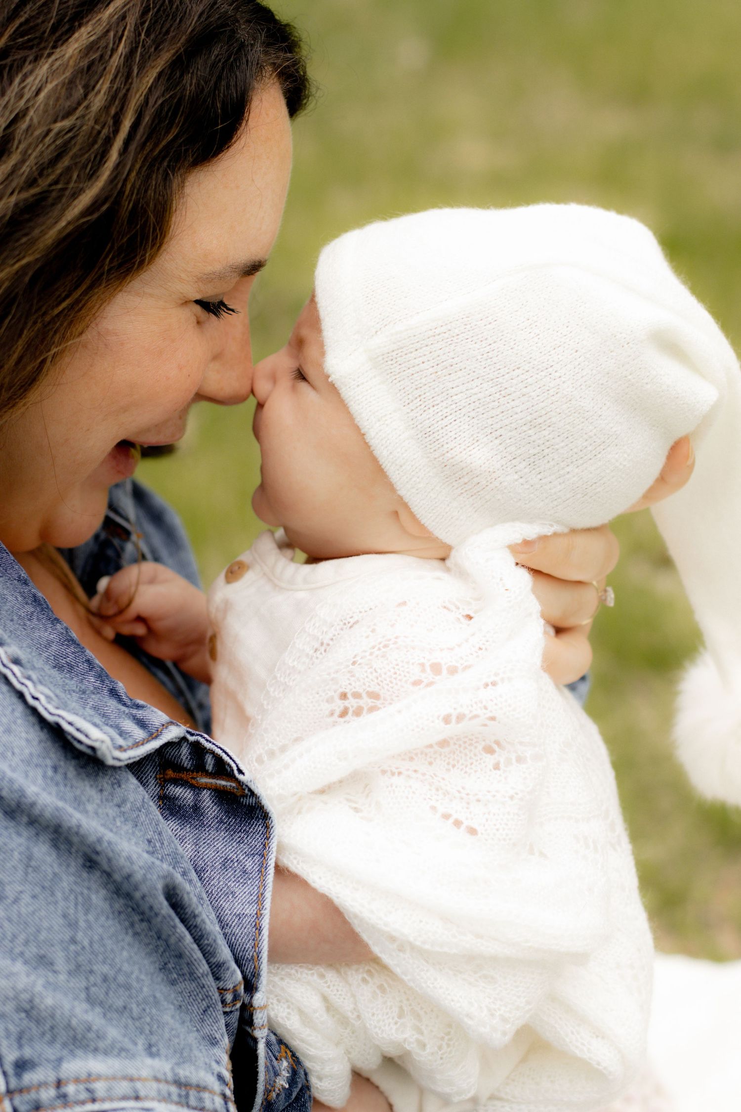 Smiling mother in denim jacket shares a nose touch moment with baby in white knit hat.