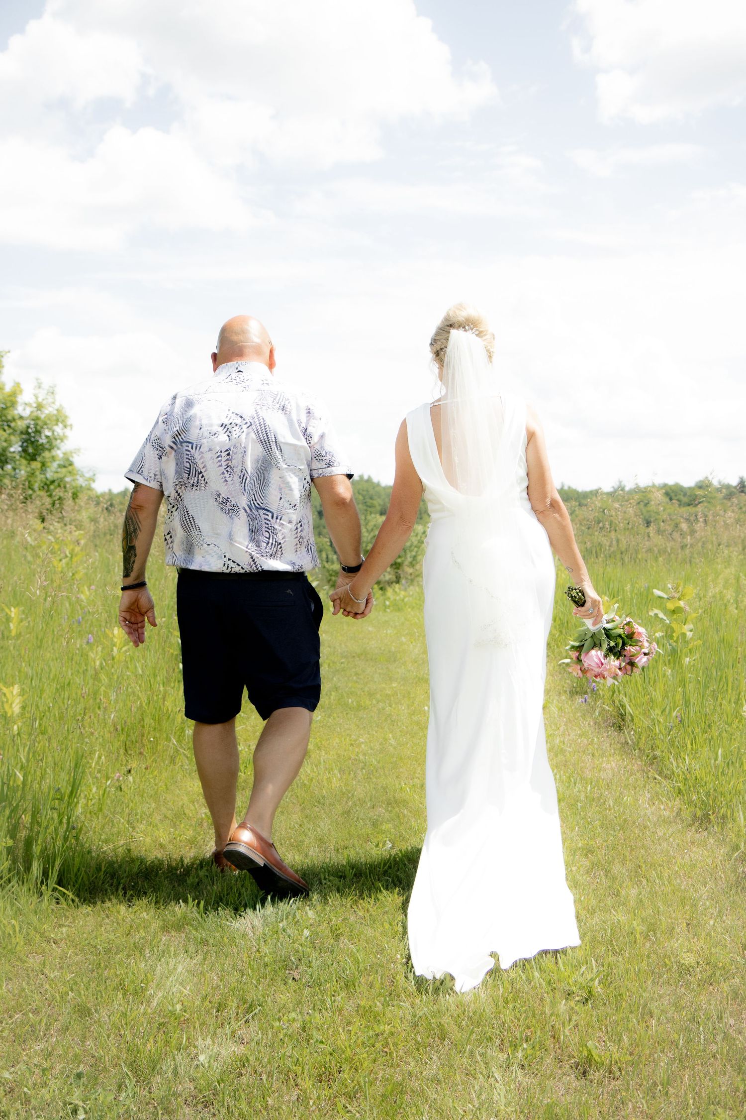 Bride and groom walking hand in hand through a green meadow, bride in white gown with veil holding pink bouquet.