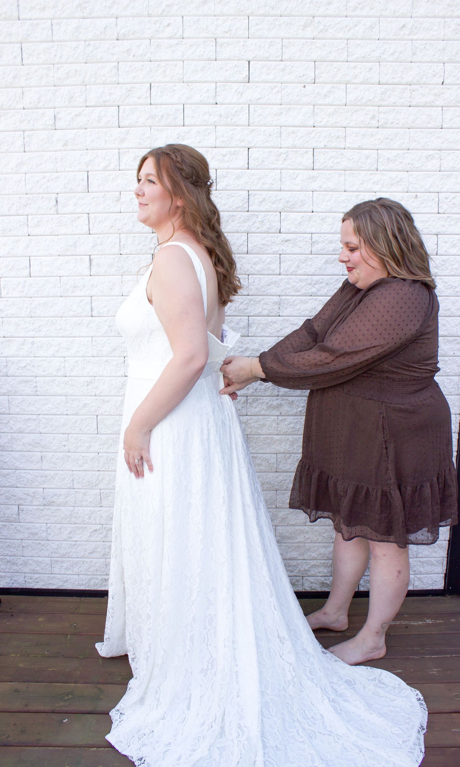 A woman in a white lace wedding gown being helped with her dress by another woman against a brick wall.