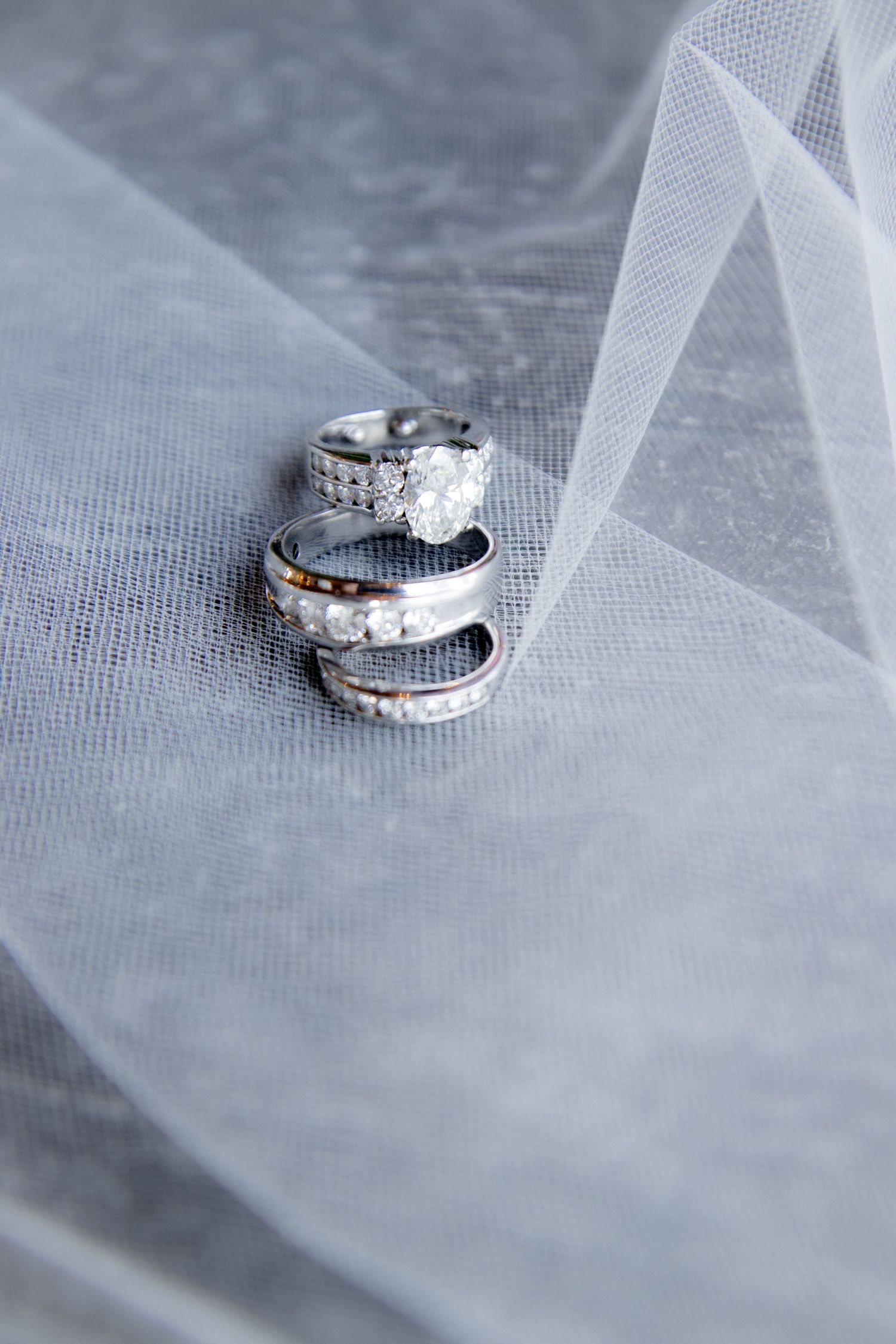 Three silver wedding rings stacked on white tulle fabric, featuring diamond engagement ring and matching bands.