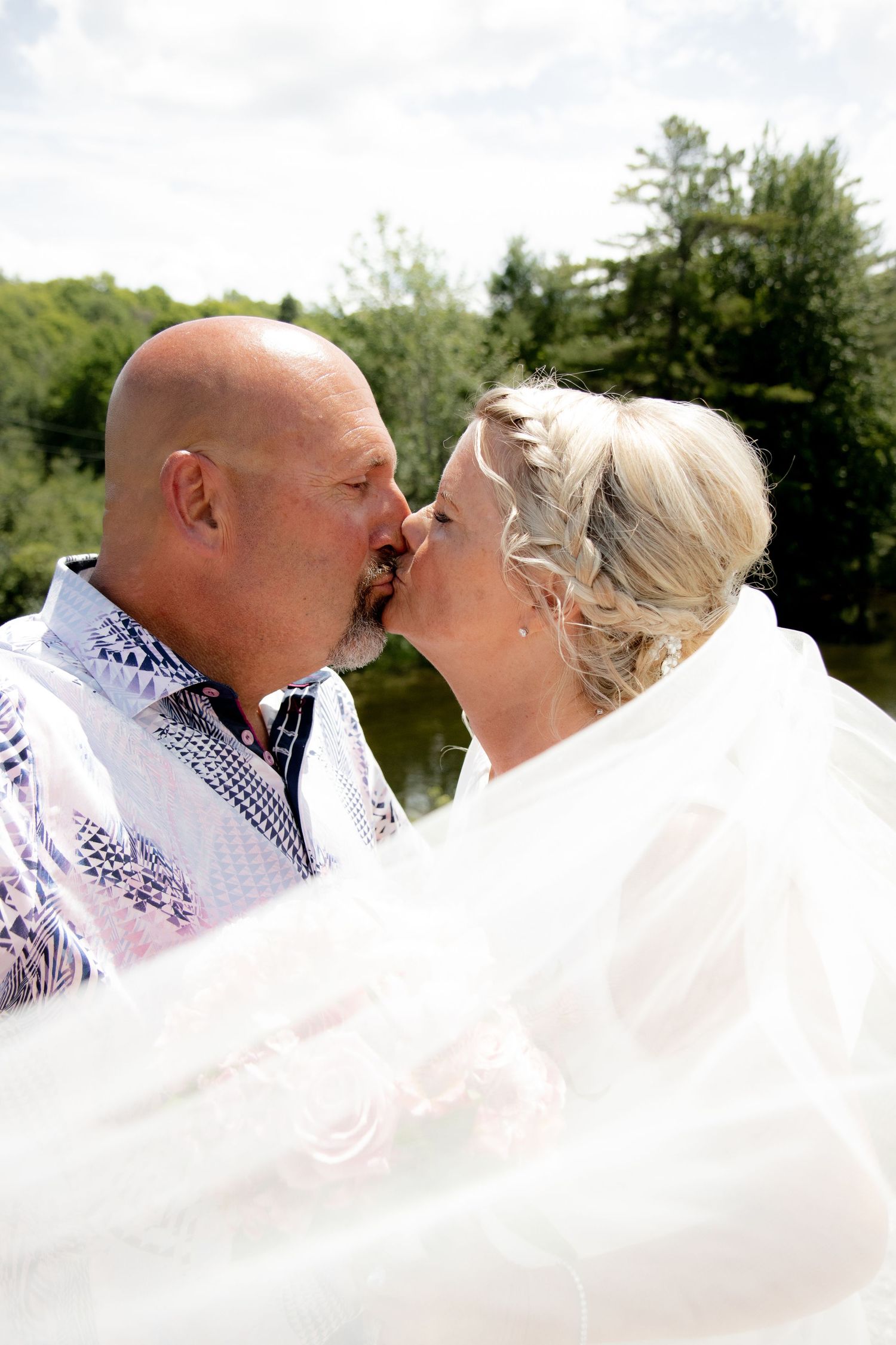 A bride and groom share a romantic kiss outdoors near a lake, with the bridal veil flowing in the breeze.