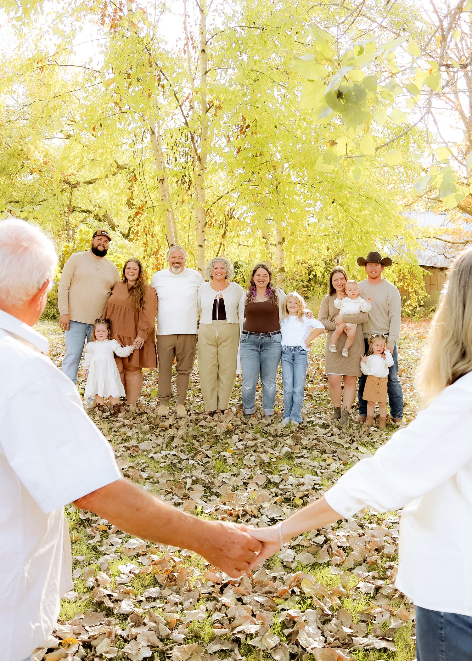 Elderly couple holds hands in foreground as multigenerational family gathers among autumn trees with golden foliage.