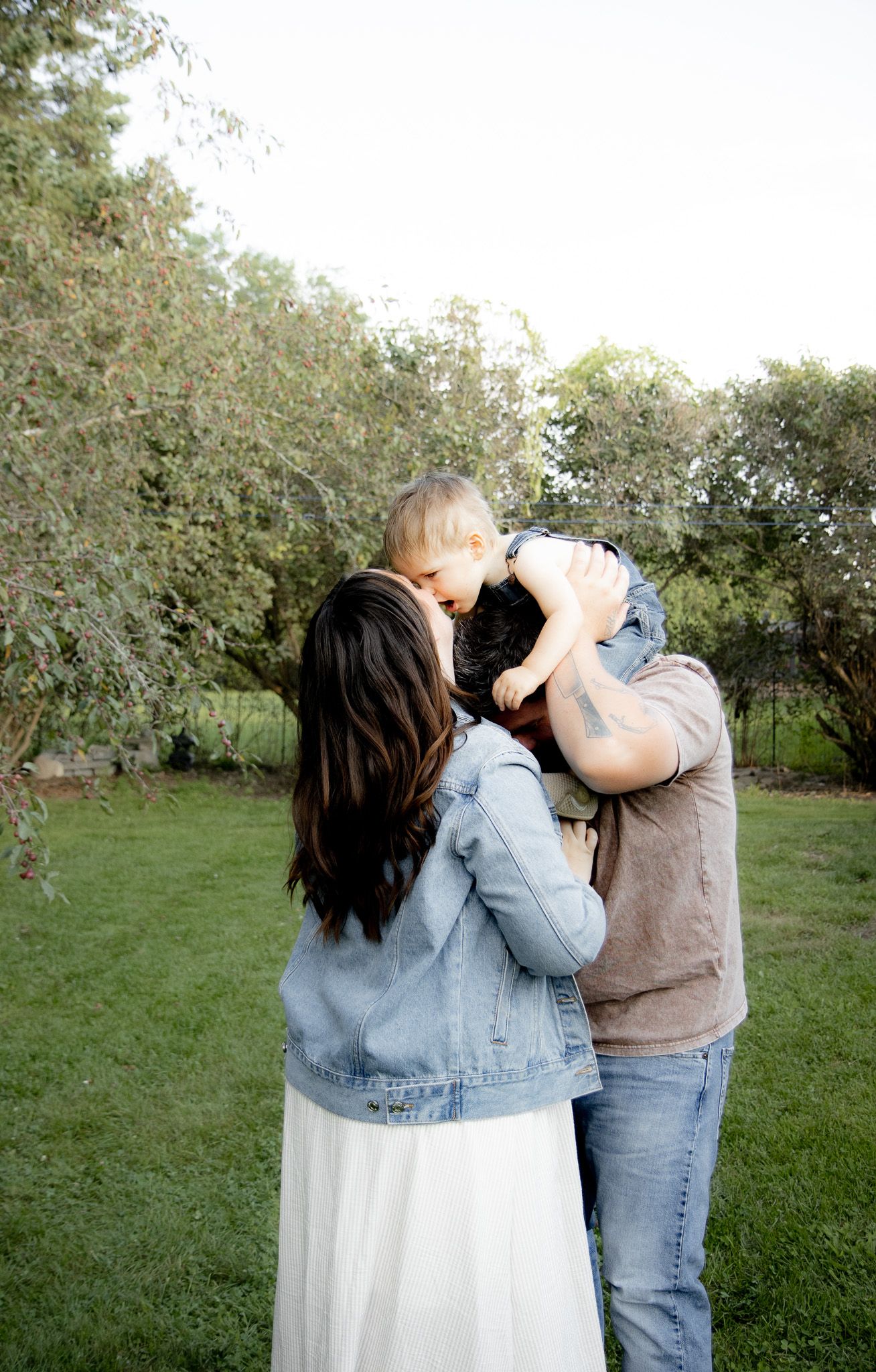 A couple shares a tender moment lifting their baby outdoors in a lush green garden setting.