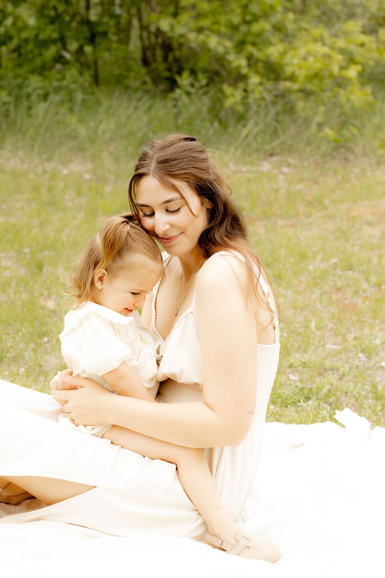 A serene mother in a white dress sits on a blanket outdoors cradling her young child in soft natural light.