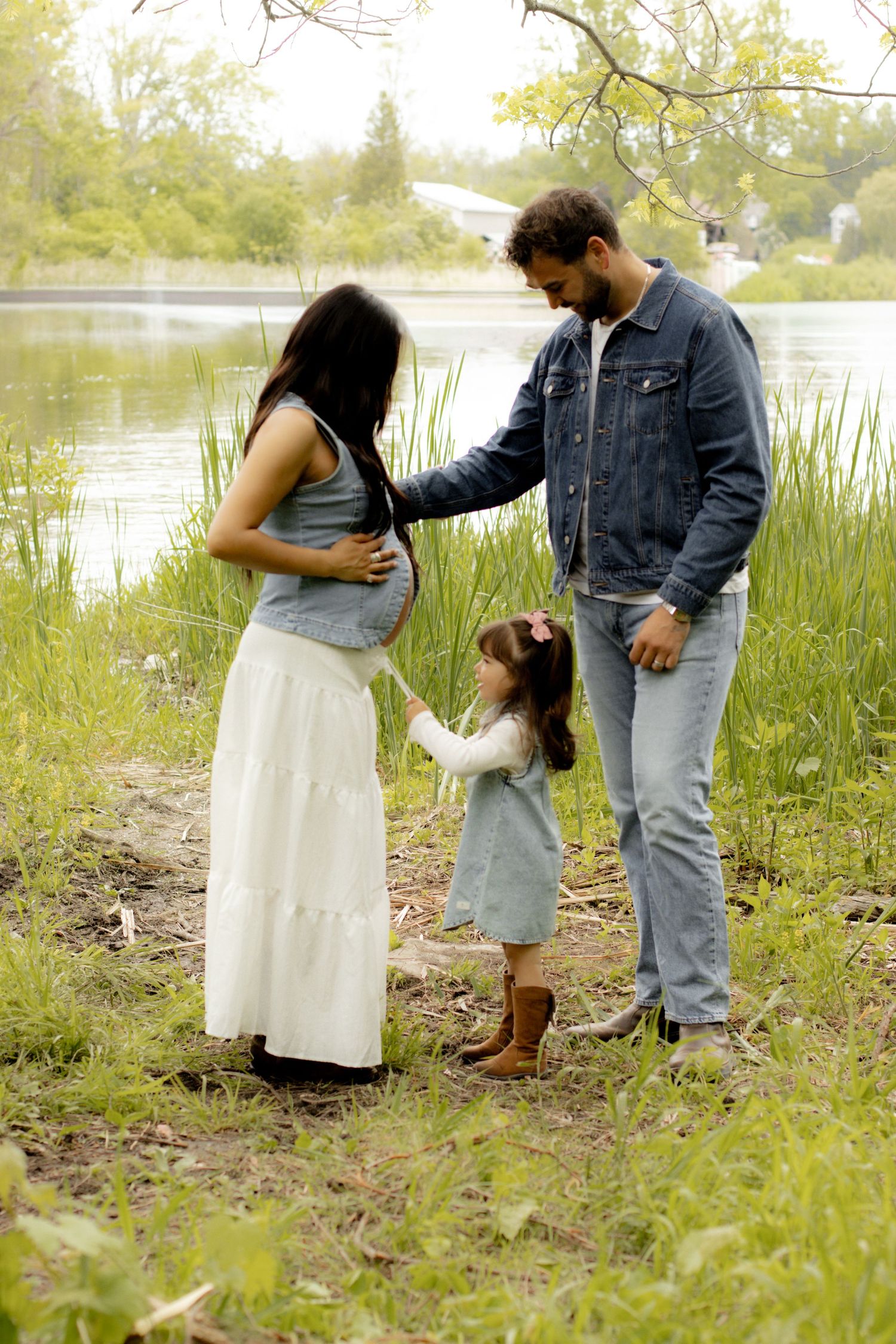 Pregnant woman with family by a lake, husband and young daughter touching her baby bump outdoors.