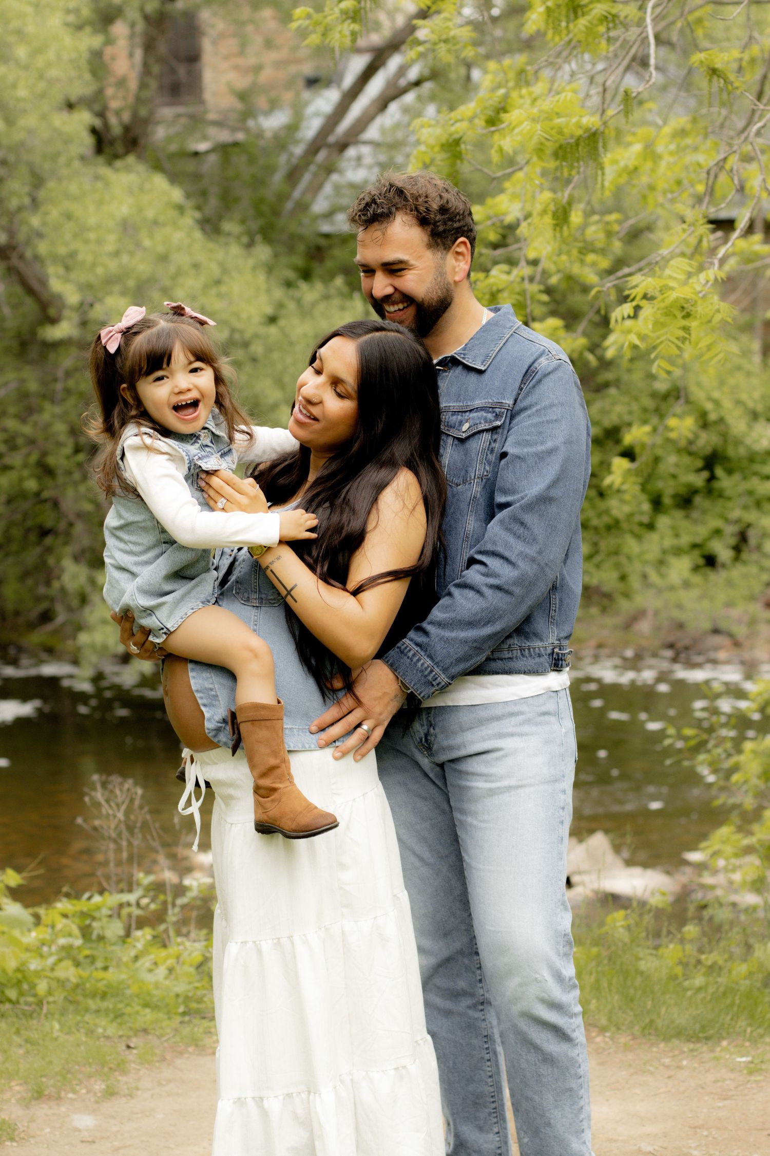 A joyful family of three stands by a creek, with parents holding their laughing toddler daughter outdoors.