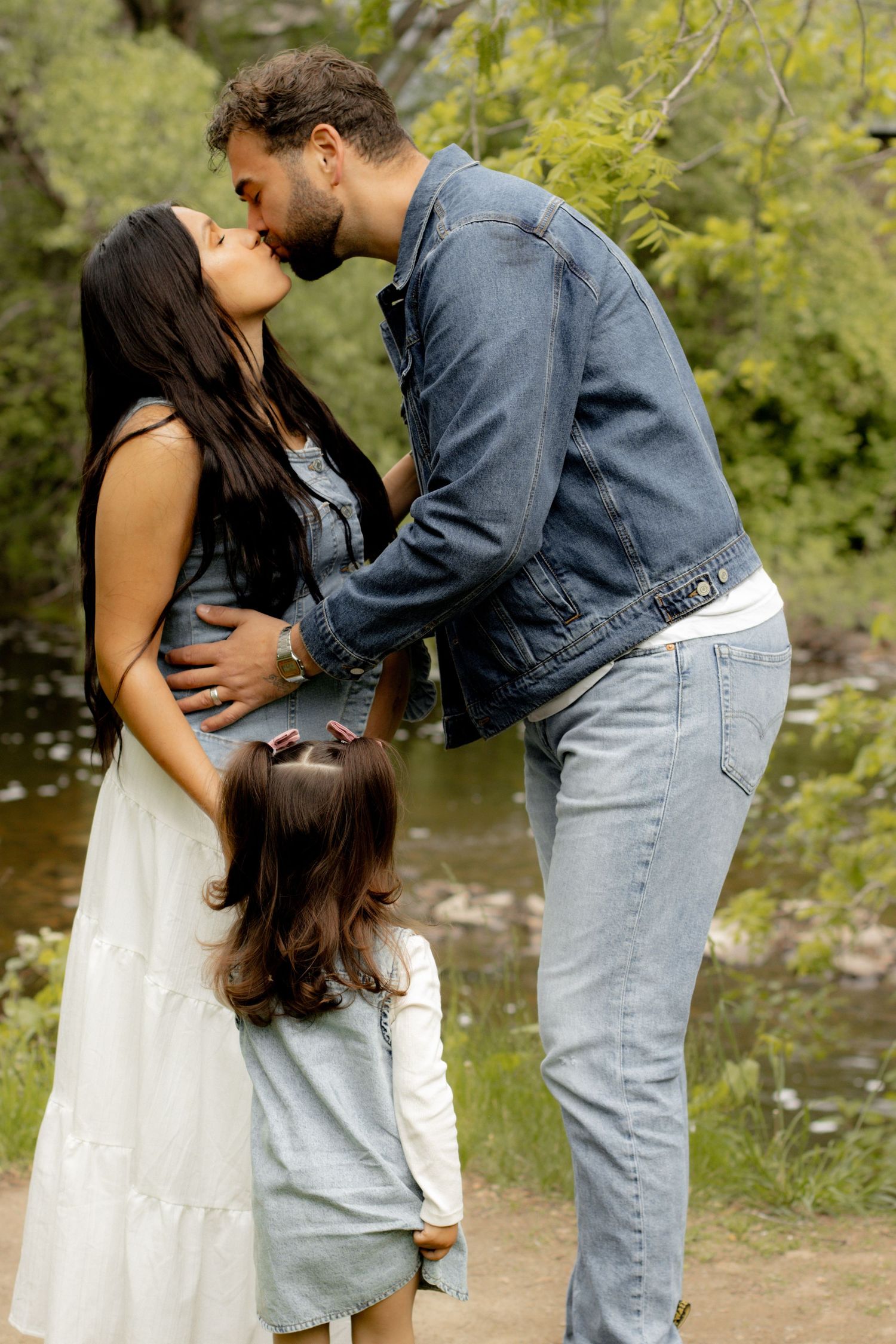 A pregnant woman and her partner share a kiss outdoors while their young daughter stands between them.