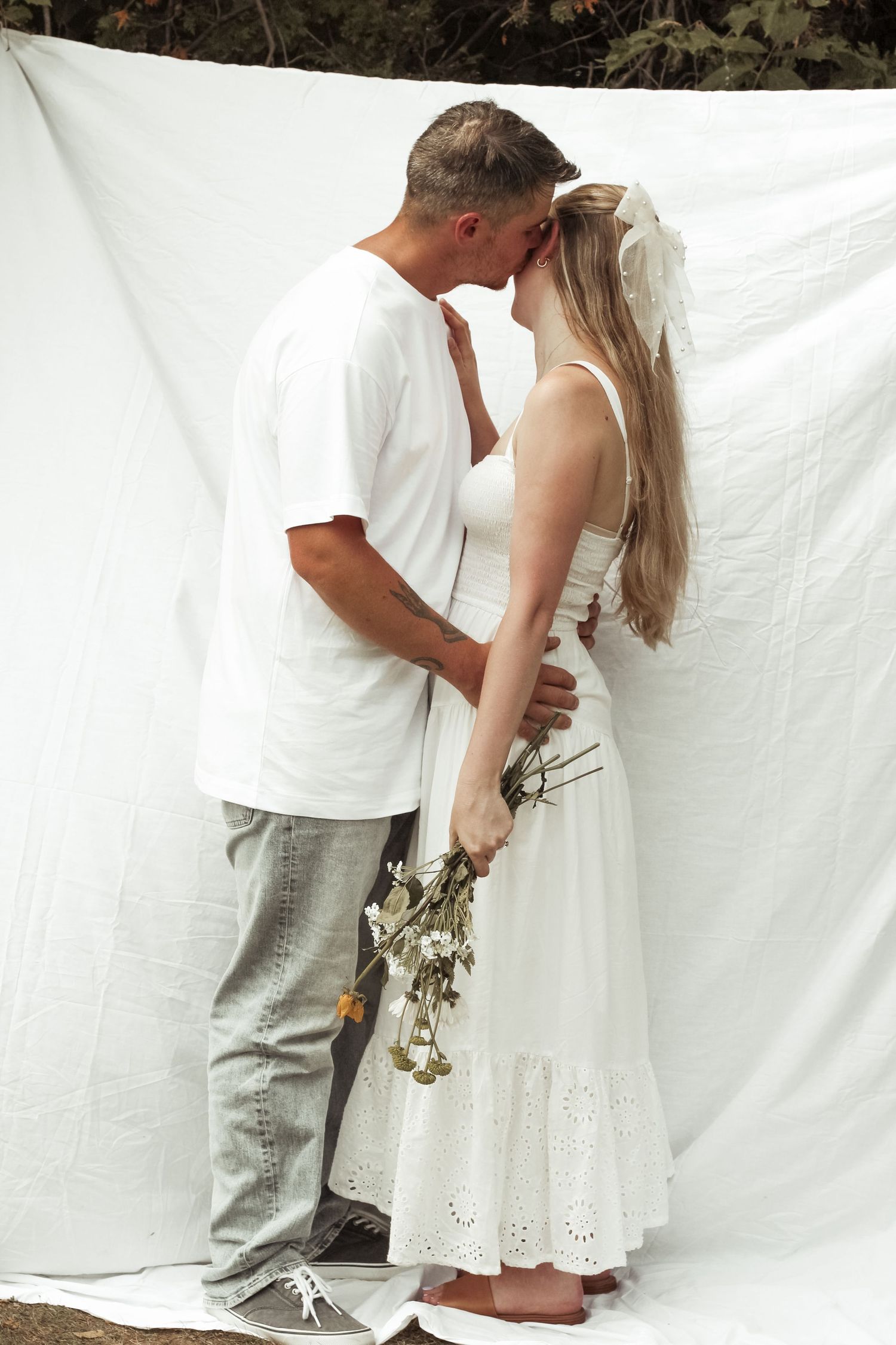 A couple shares a romantic kiss in front of a white backdrop, the bride holding a wildflower bouquet in a white dress.