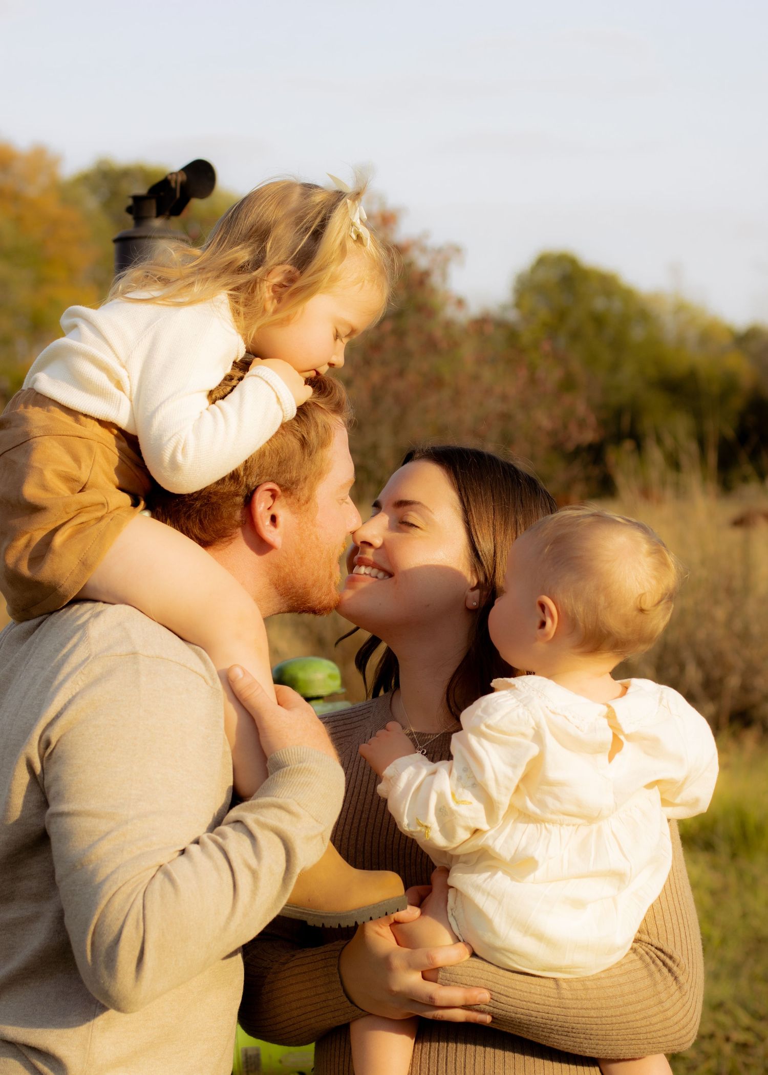 Happy parents share a tender moment while holding two young children outdoors in warm golden autumn light.