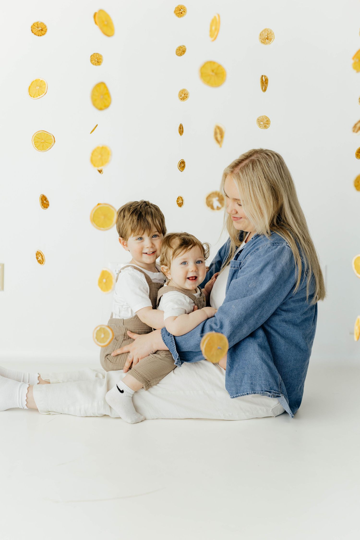 Blonde mother sitting on floor with two young children under falling lemon slice decorations.