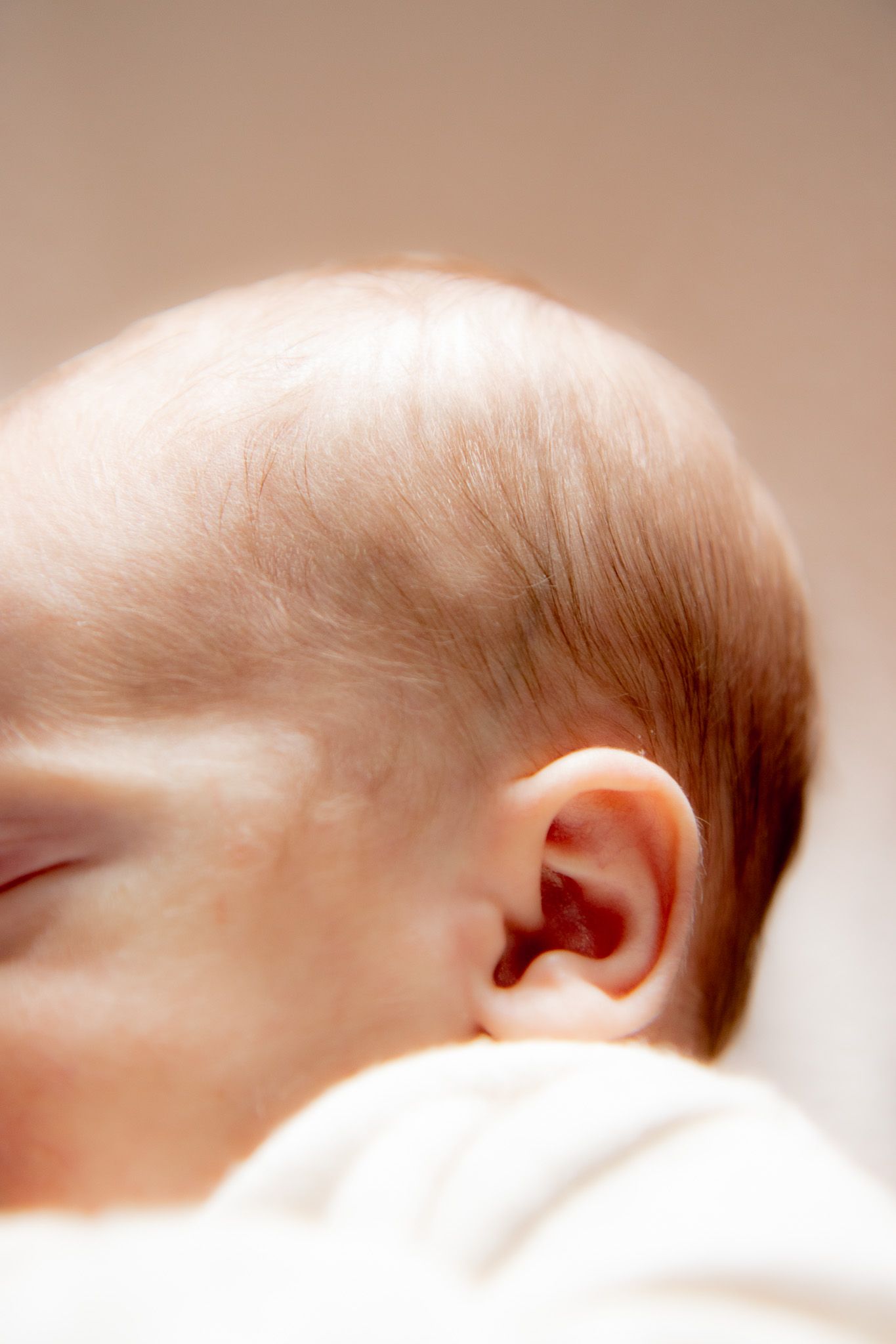 Close-up side profile of a sleeping newborn baby with fine reddish hair and a delicate ear against a soft background.