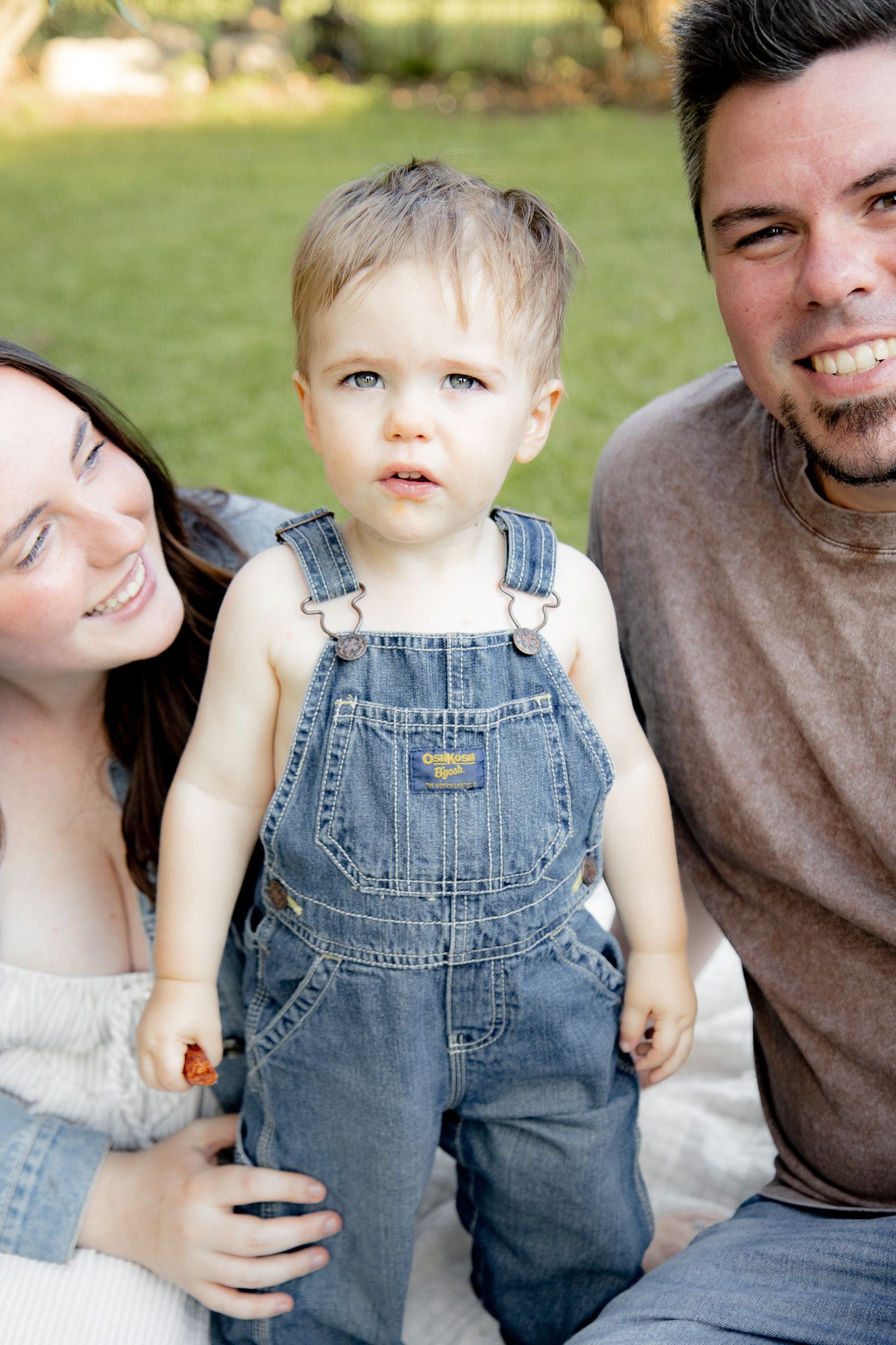 Toddler boy in denim overalls stands between smiling parents outdoors on green grass.
