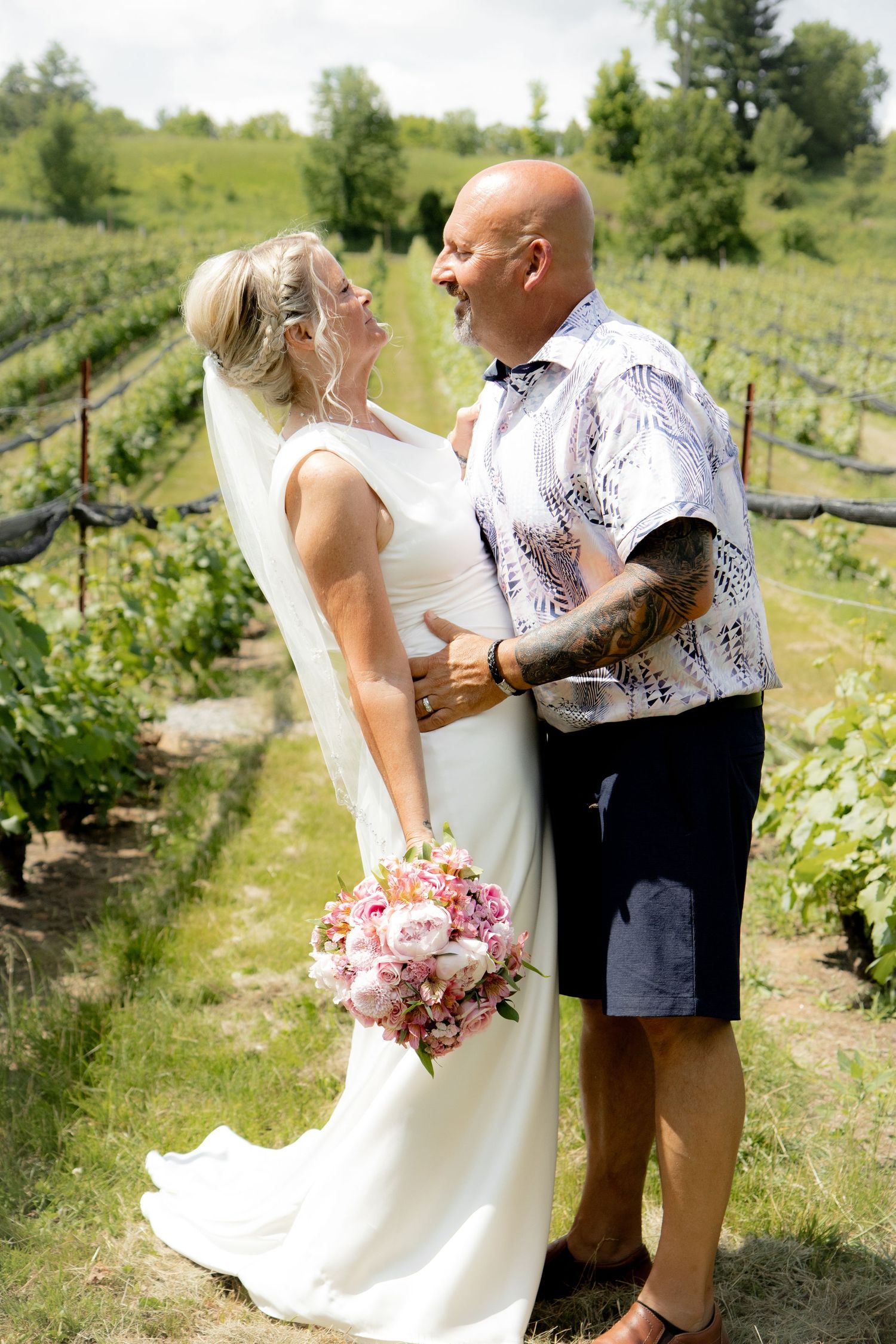Bride and groom share a joyful moment in a vineyard, bride holding pink bouquet in white gown, groom in floral shirt.