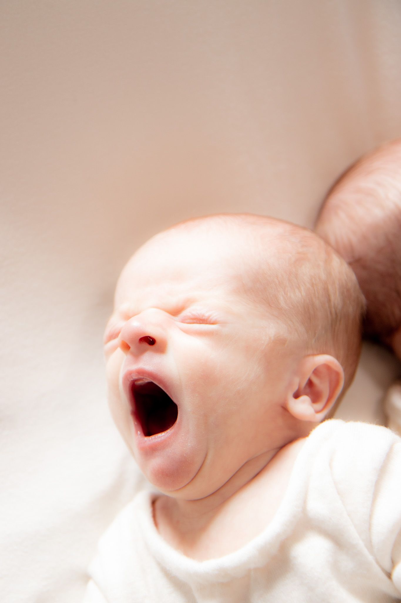 Close-up of a newborn baby yawning with eyes closed, wearing a white onesie, lying on a soft surface.