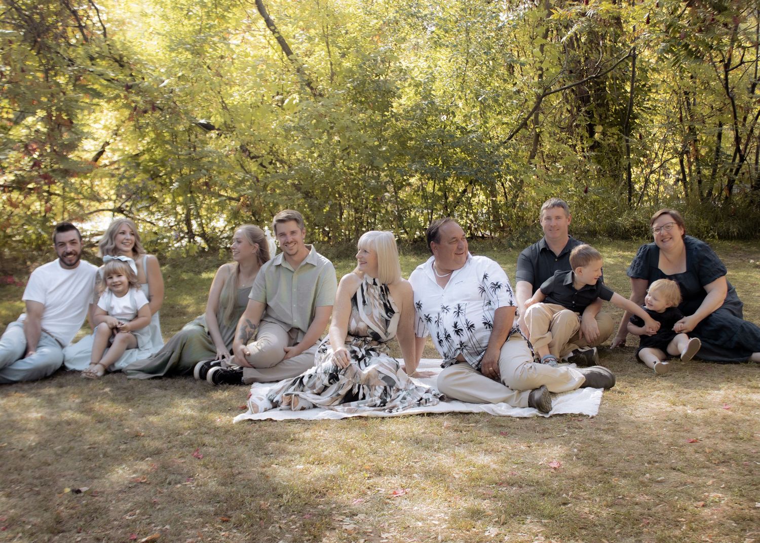 Extended family group sitting together on grass in wooded park during warm autumn day.