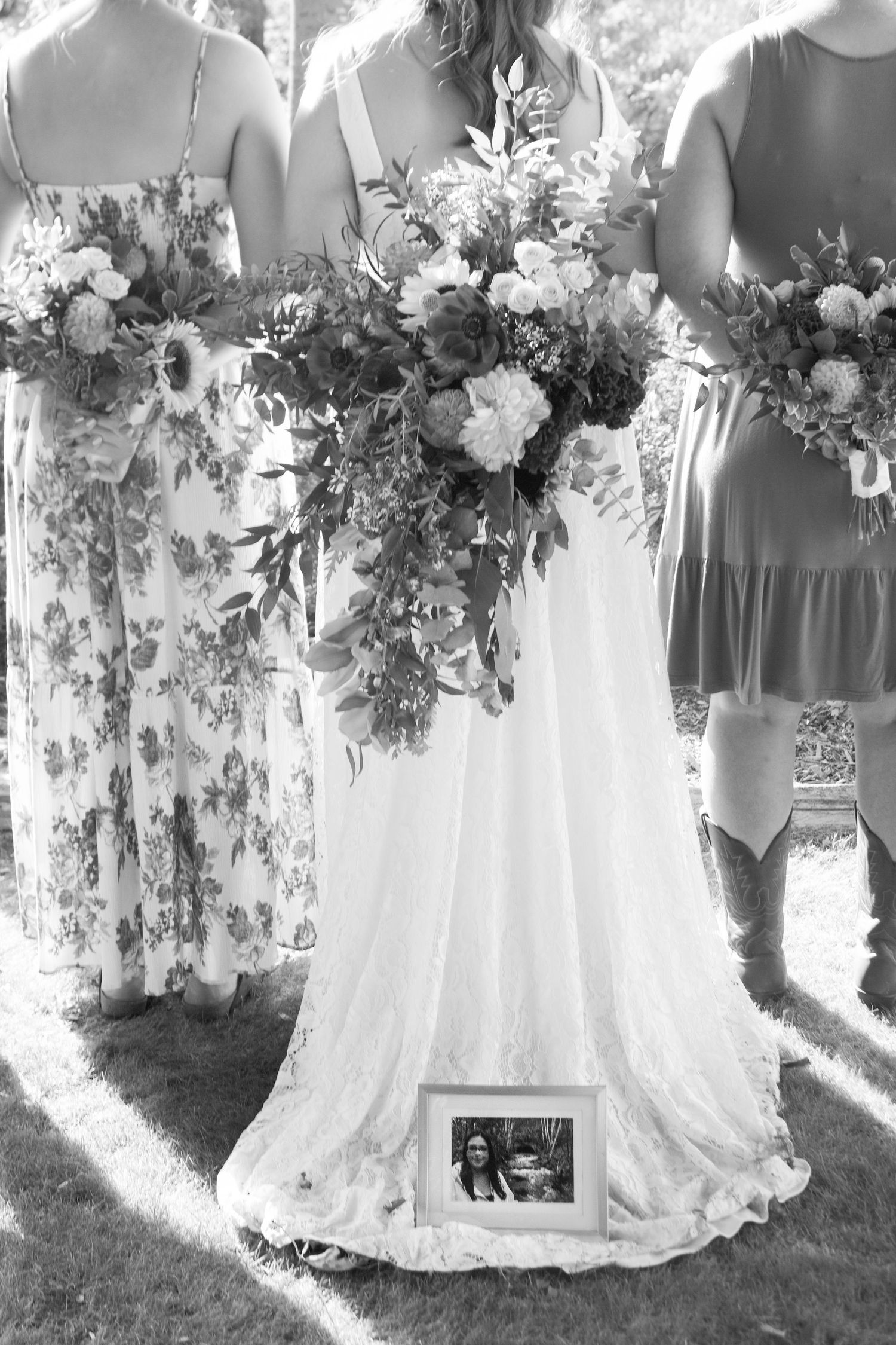 Black and white wedding photo of bride holding floral bouquet with memorial photo tucked in dress hem, flanked by bridesmaids.