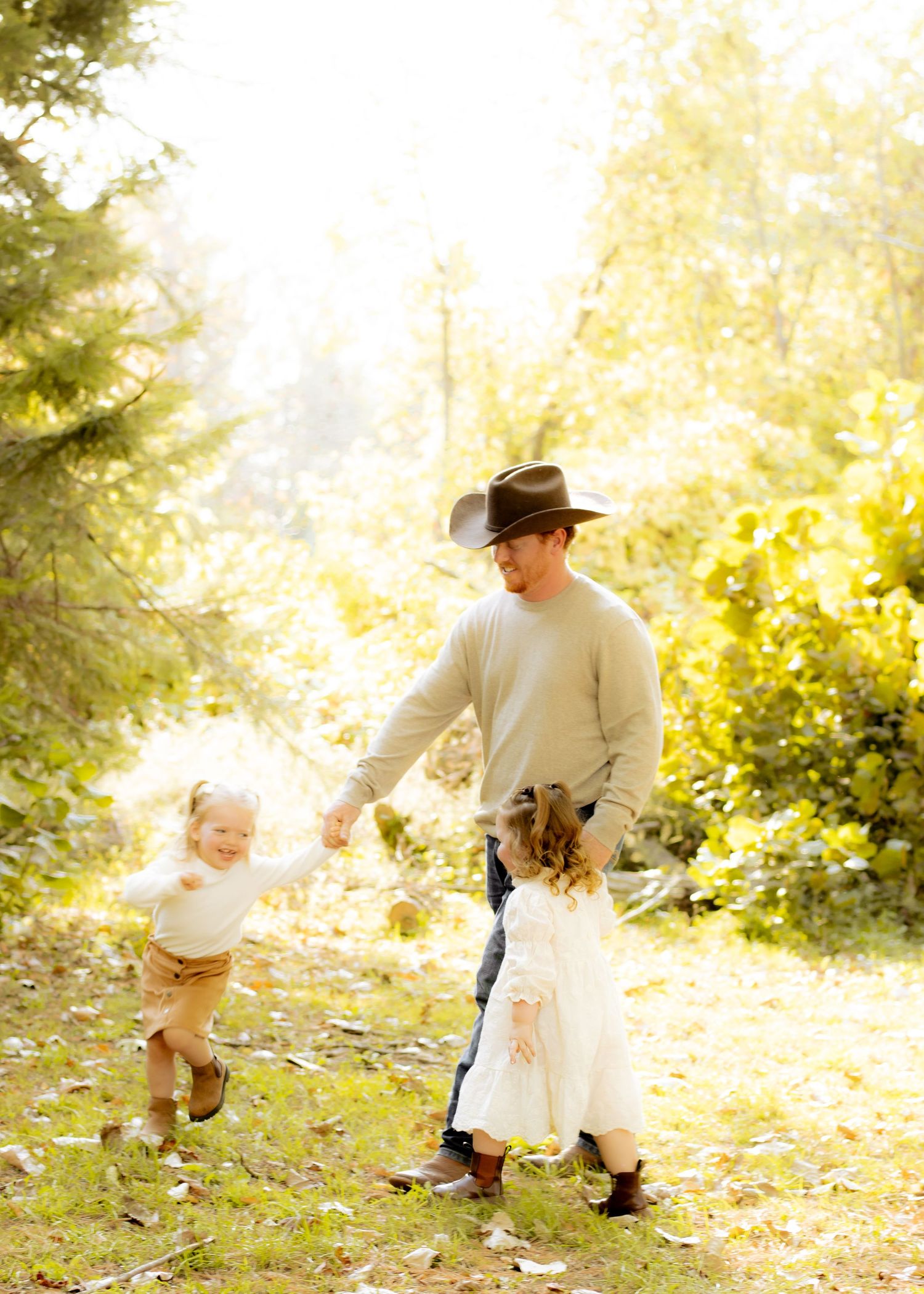Father in cowboy hat dances with two young children in sunlit autumn forest clearing with golden trees.