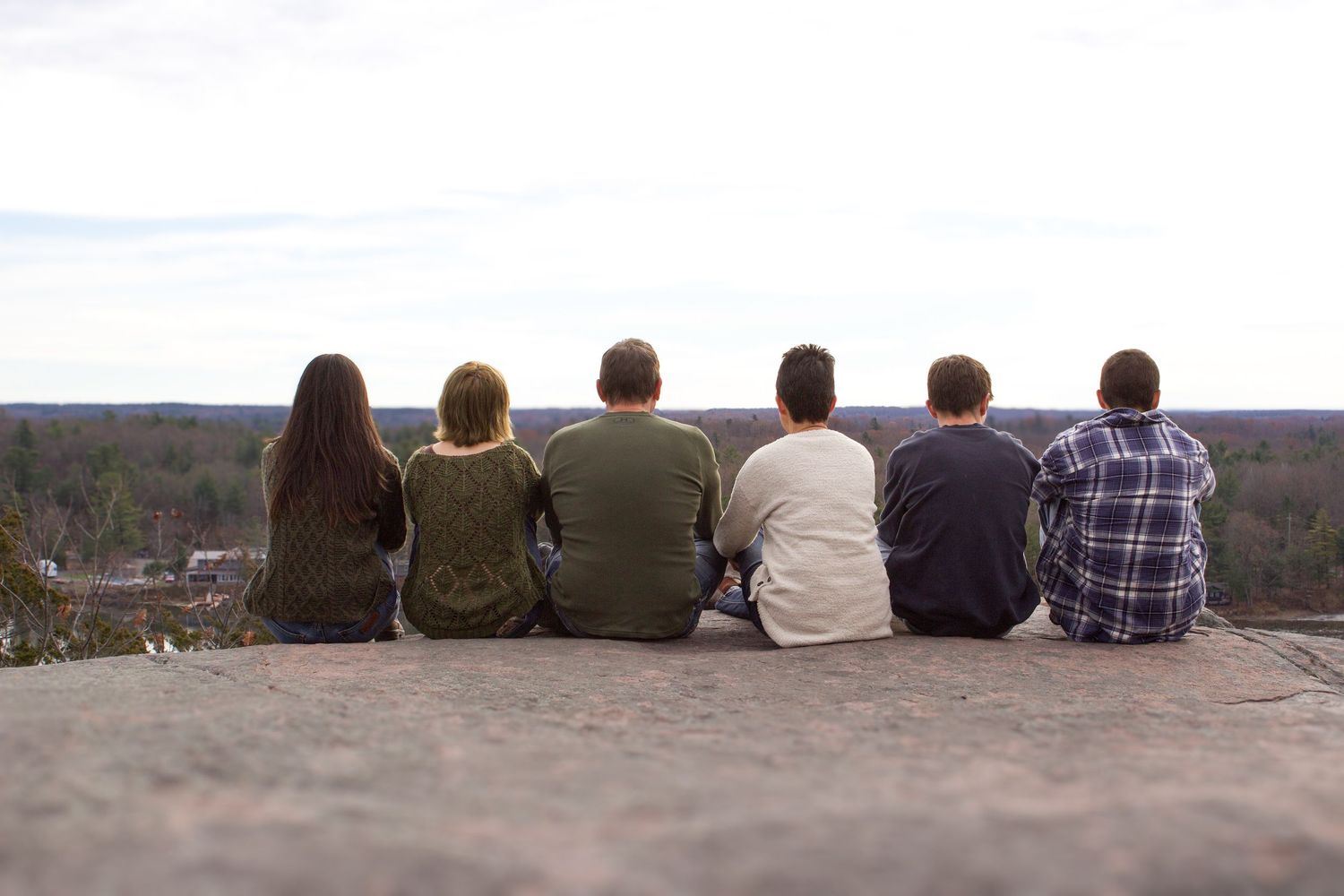 Six family members sit with backs to camera on rocky ledge, overlooking vast autumn forest landscape.