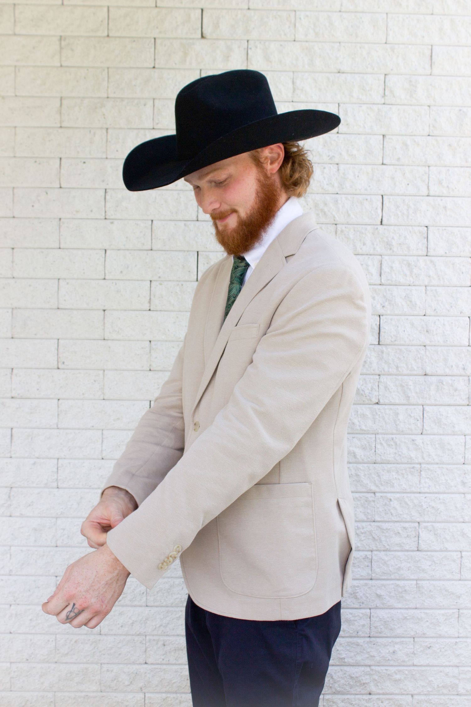 A bearded groom in a tan blazer and black cowboy hat adjusting his sleeve against a white brick wall.