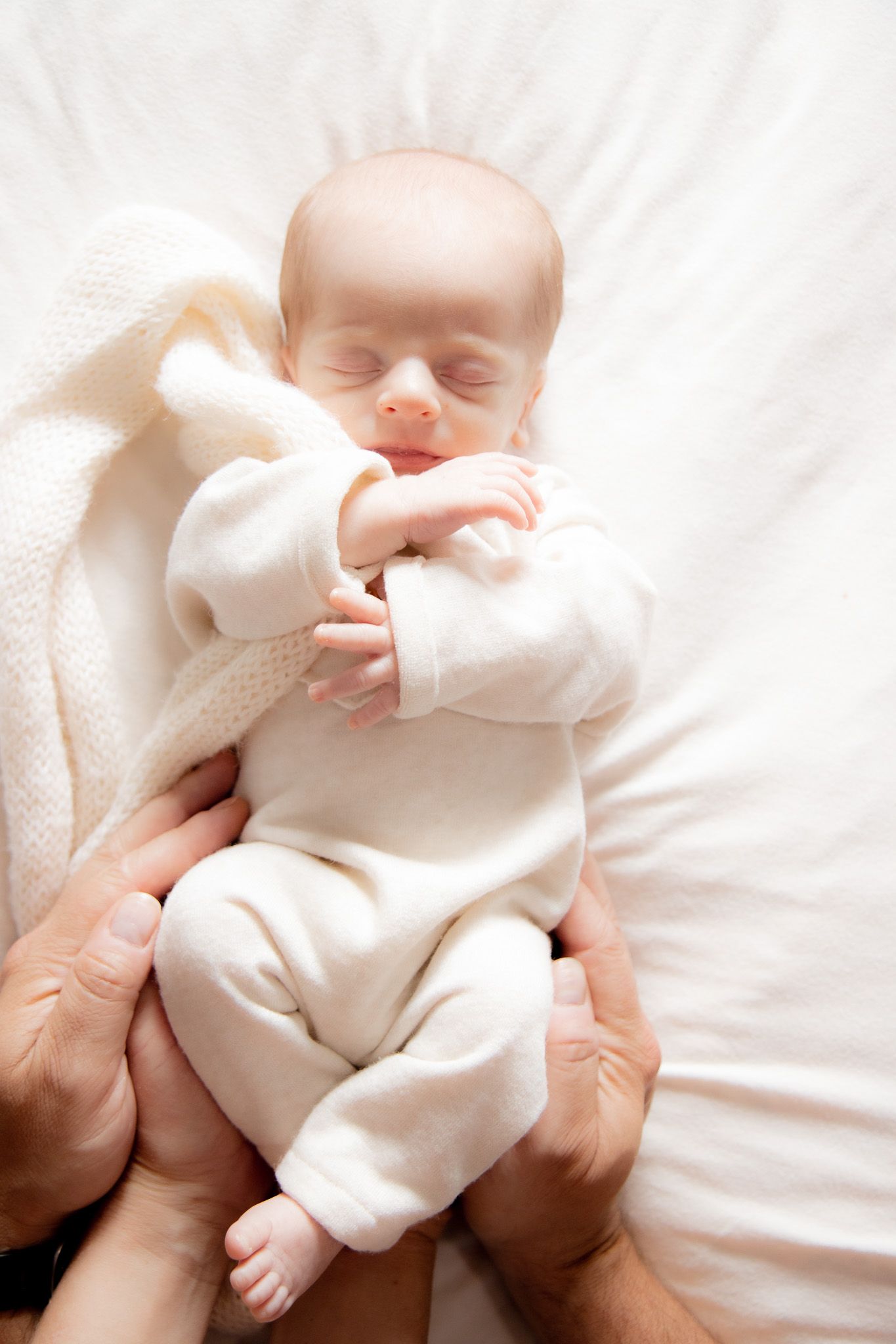 Newborn baby sleeping peacefully in white onesie cradled in parents hands on soft white blanket.