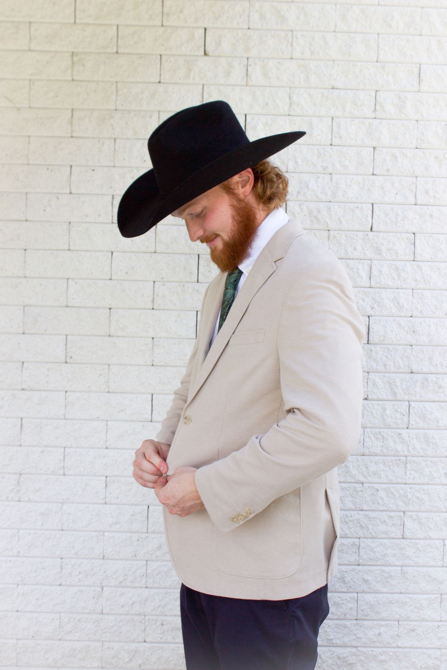 Groom in tan suit and black cowboy hat buttoning jacket against white brick wall.