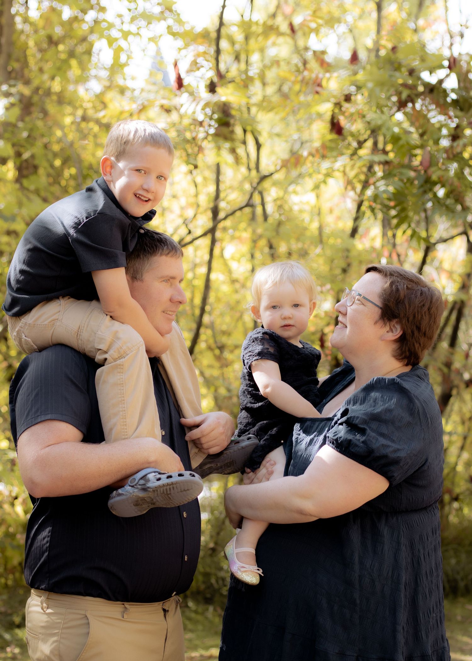 Young family poses outdoors in fall, dad holding son on shoulders, mom cradling toddler, surrounded by autumn foliage.