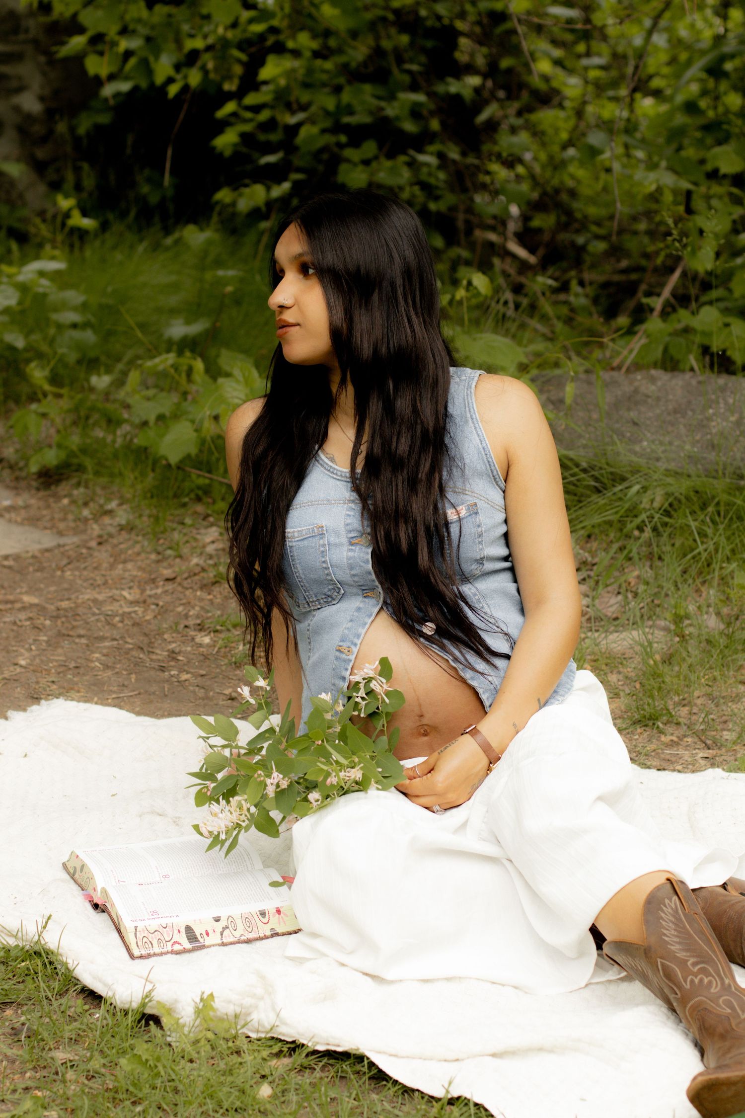 Pregnant woman with long dark hair sitting on white blanket outdoors with flowers and book.