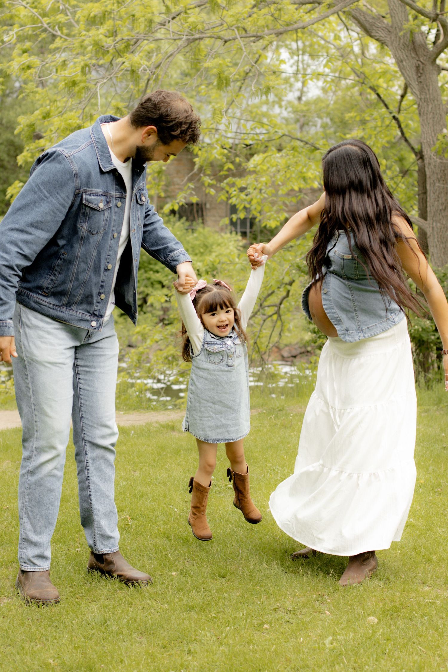 A joyful family swinging their toddler girl between them in a lush green park, all wearing denim outfits.