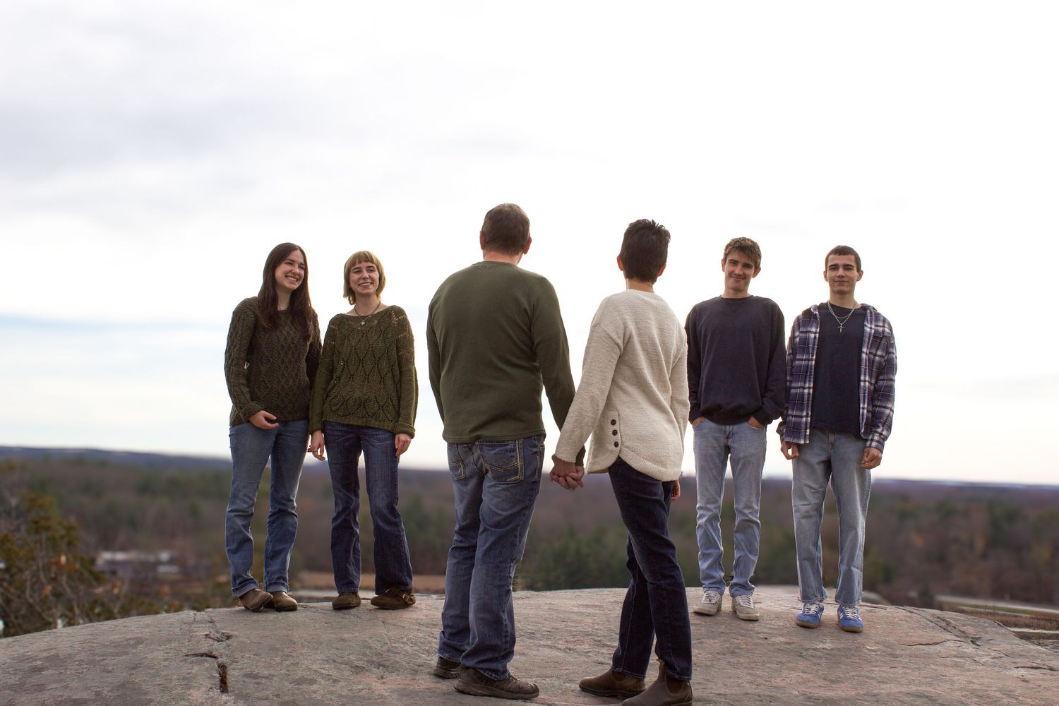 Family of six stands on rocky overlook with expansive forest landscape, two members holding hands in center.