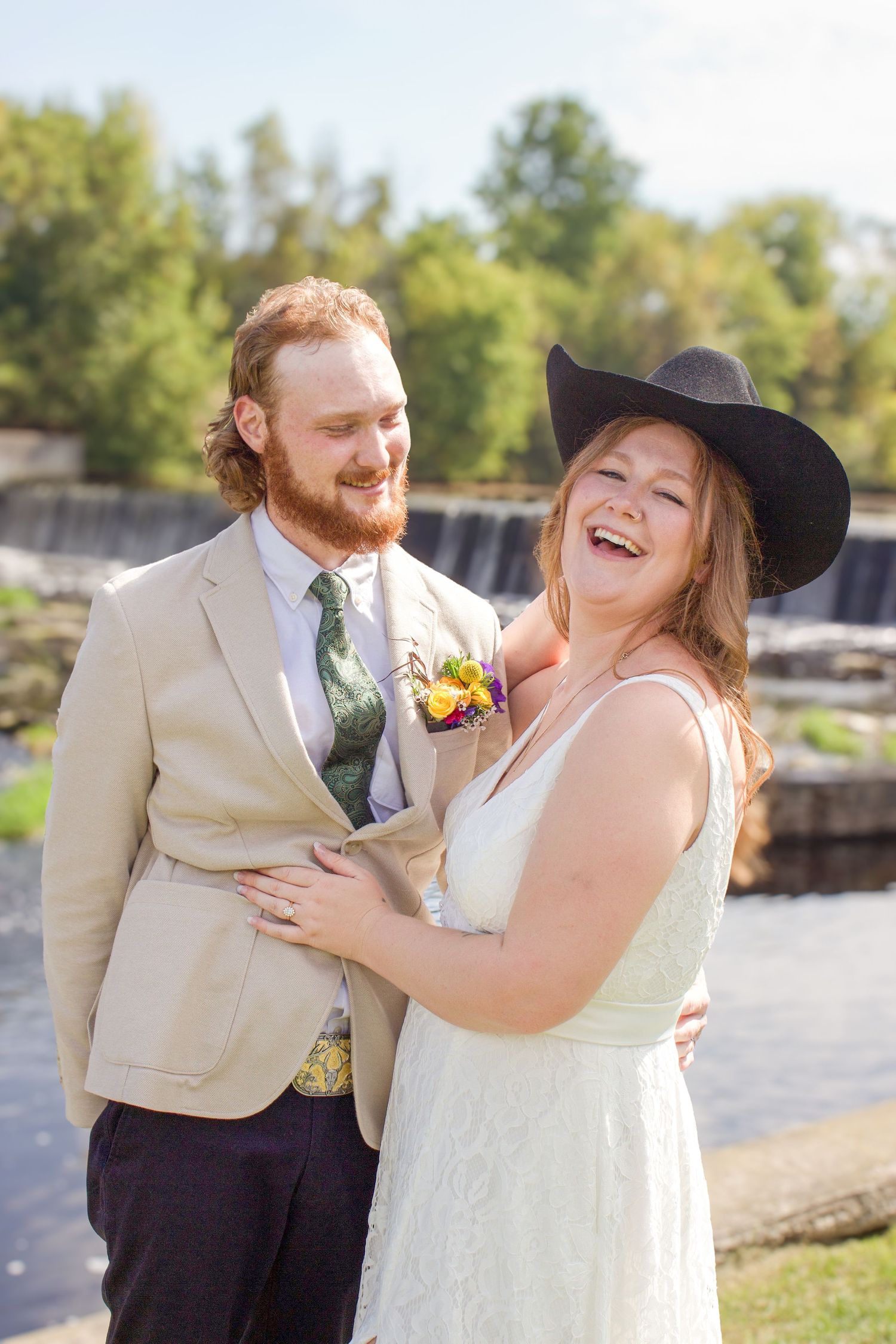 Laughing bride in cowboy hat embraces bearded groom in tan suit near waterfall.