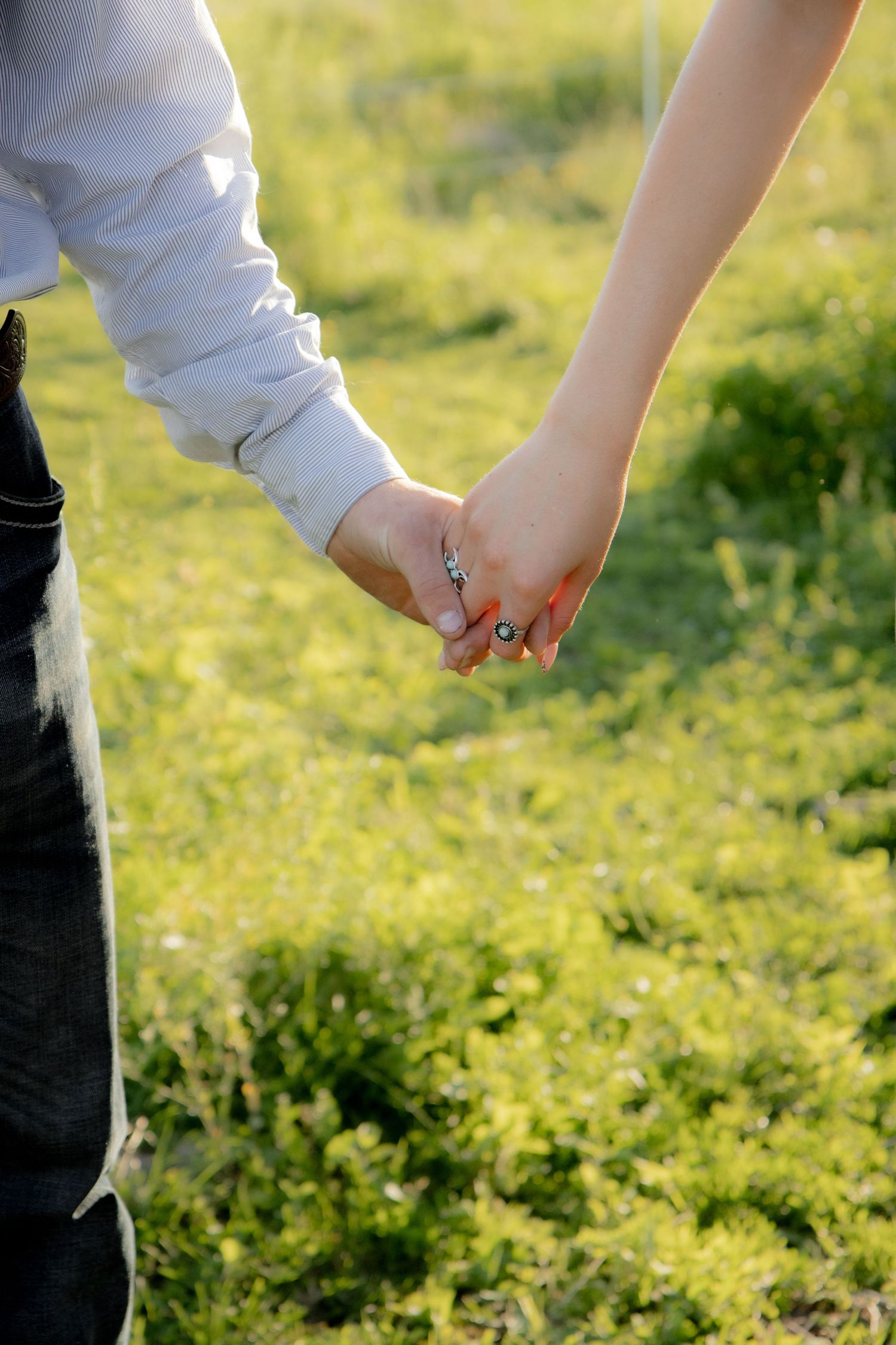 Couple holding hands with engagement ring on finger, walking through lush green garden in sunlight.