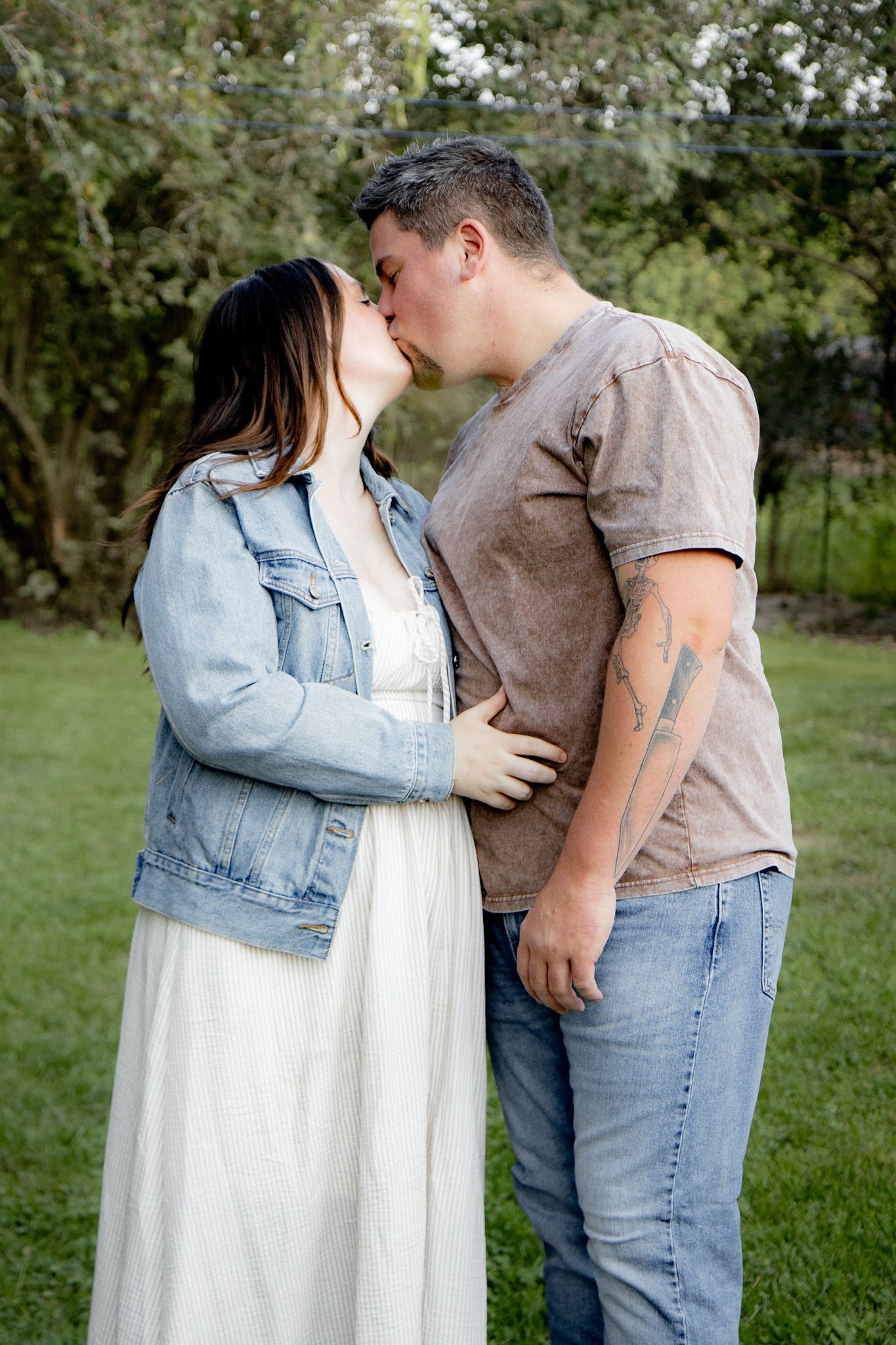 A couple shares a romantic kiss outdoors, woman in white dress and denim jacket, man with tattoo in brown shirt.