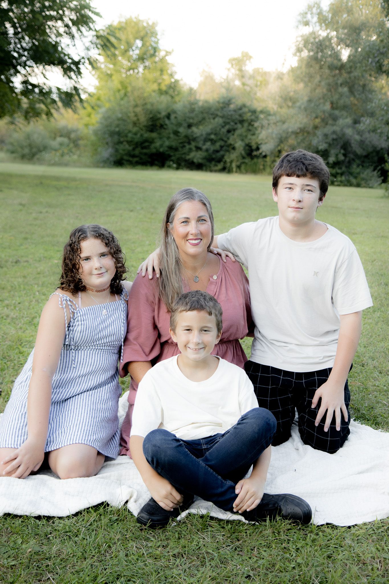 A smiling family of four poses outdoors on a white blanket in a lush green park during golden hour.