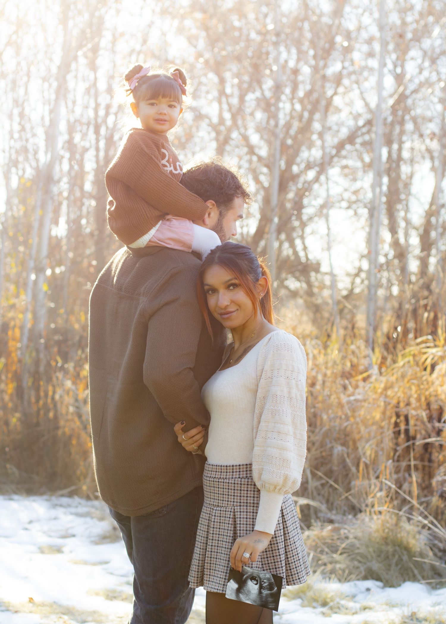 Family portrait with father carrying toddler on shoulders while mother embraces him in golden autumn light.