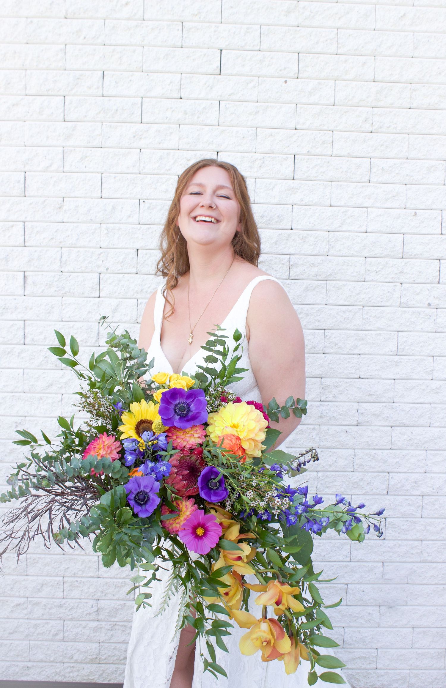 A smiling bride in a white dress holding a large colorful bouquet of purple, yellow, and pink flowers.